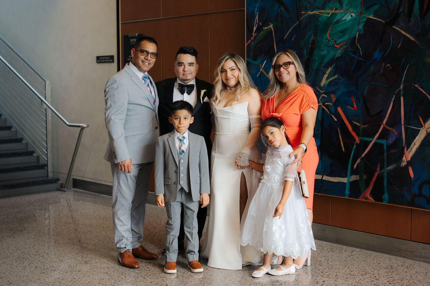 A wedding party stands together for photos in front of a modern interior wall with colorful abstract artwork.