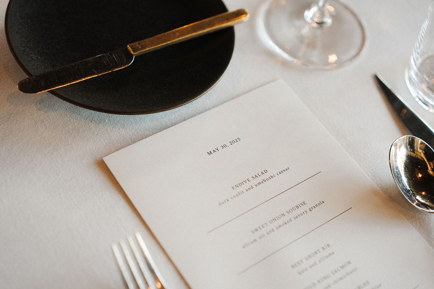 A minimalist table setting with a white menu card, chopsticks on a black plate, and wine glass on white tablecloth.