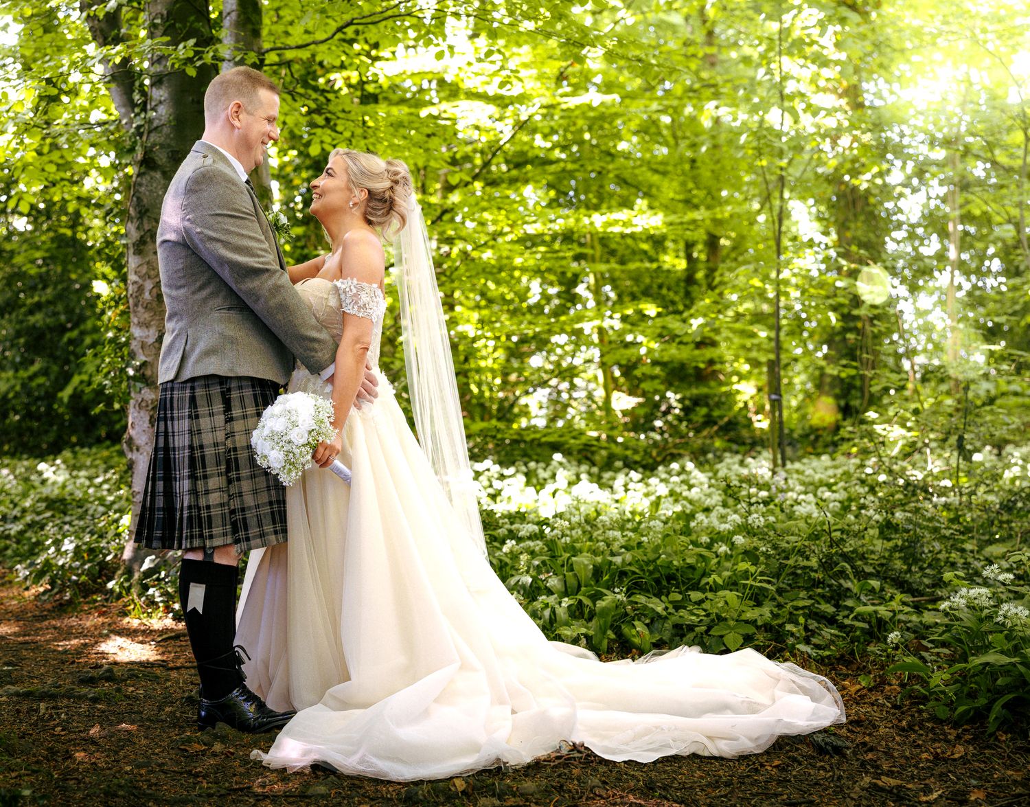 A couple shares an intimate embrace in a lush forest setting with sunlight streaming through the trees and greenery.