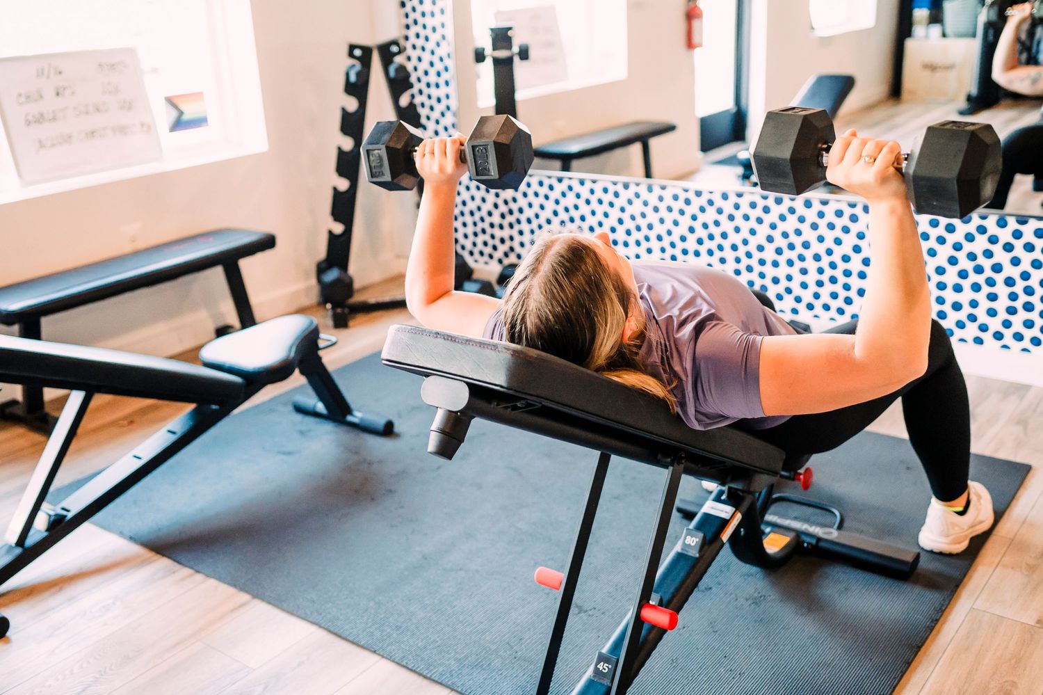 Person performs chest press workout with dumbbells while lying on bench in modern fitness studio.