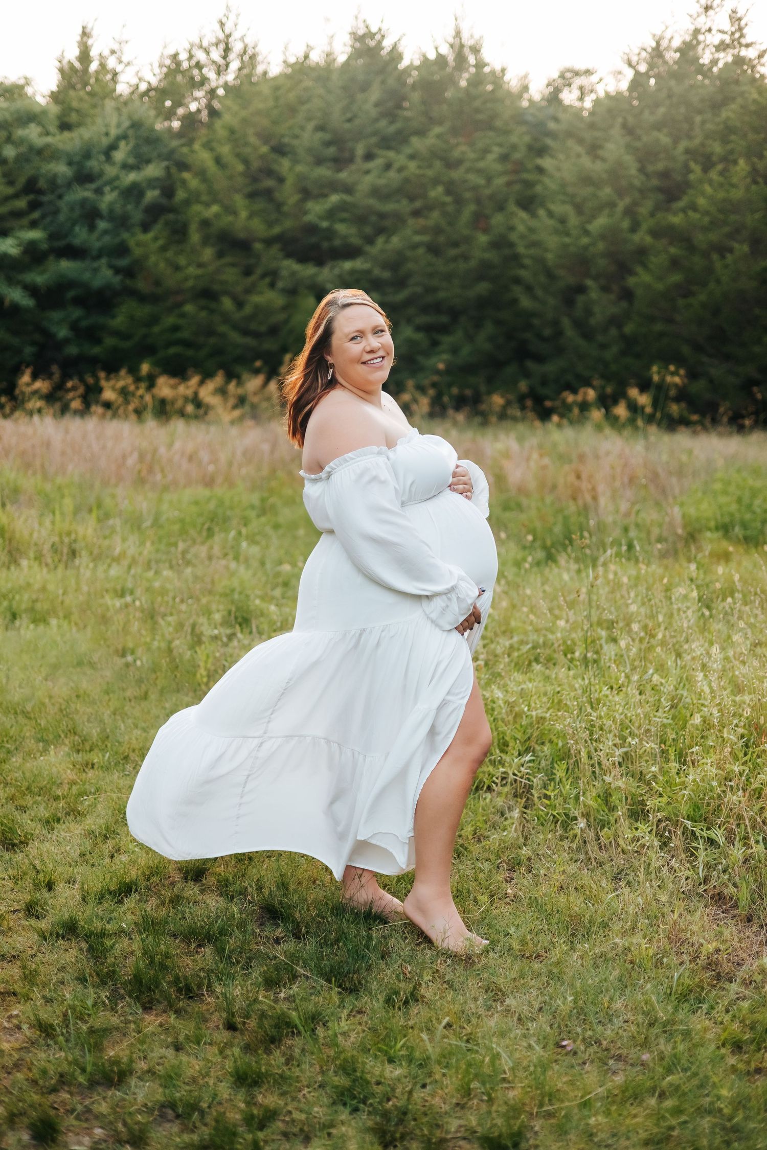 Woman in flowing white dress twirling gracefully in sunlit meadow during outdoor photo session.