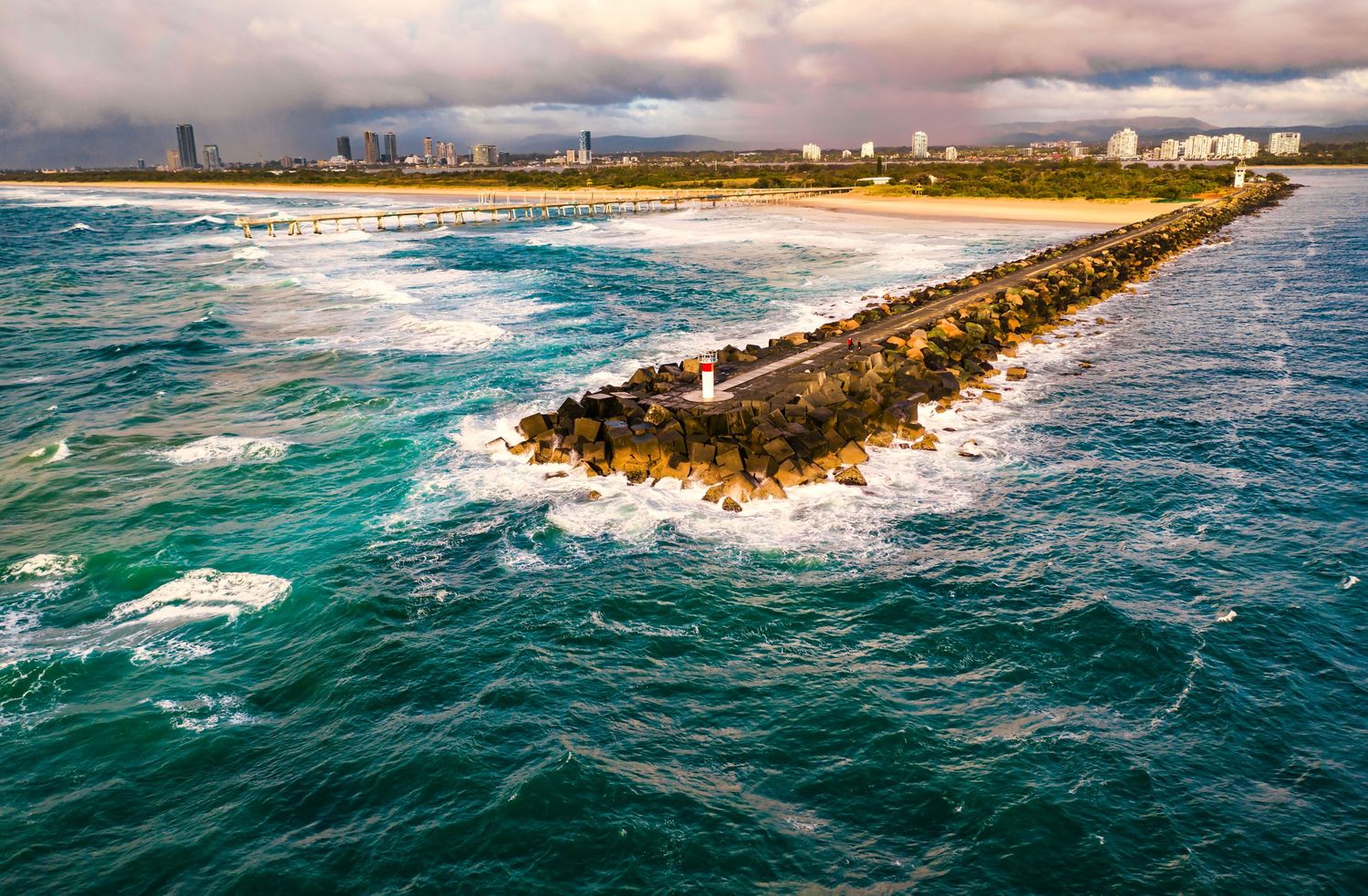 Aerial view of a rocky jetty extending into turquoise ocean waters with city skyline in background.