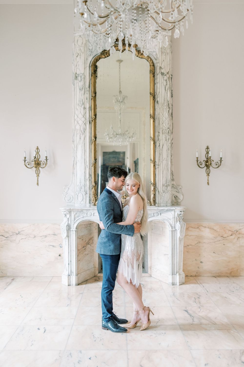 Elegant couple stands in front of ornate gilded mirror and crystal chandelier in luxurious white marble room with wall sconces.
