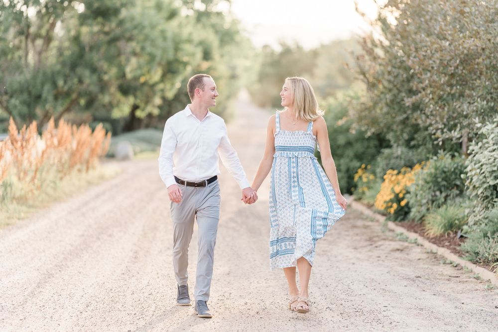 A couple walks hand in hand on a gravel path surrounded by lush greenery and summer blooms.