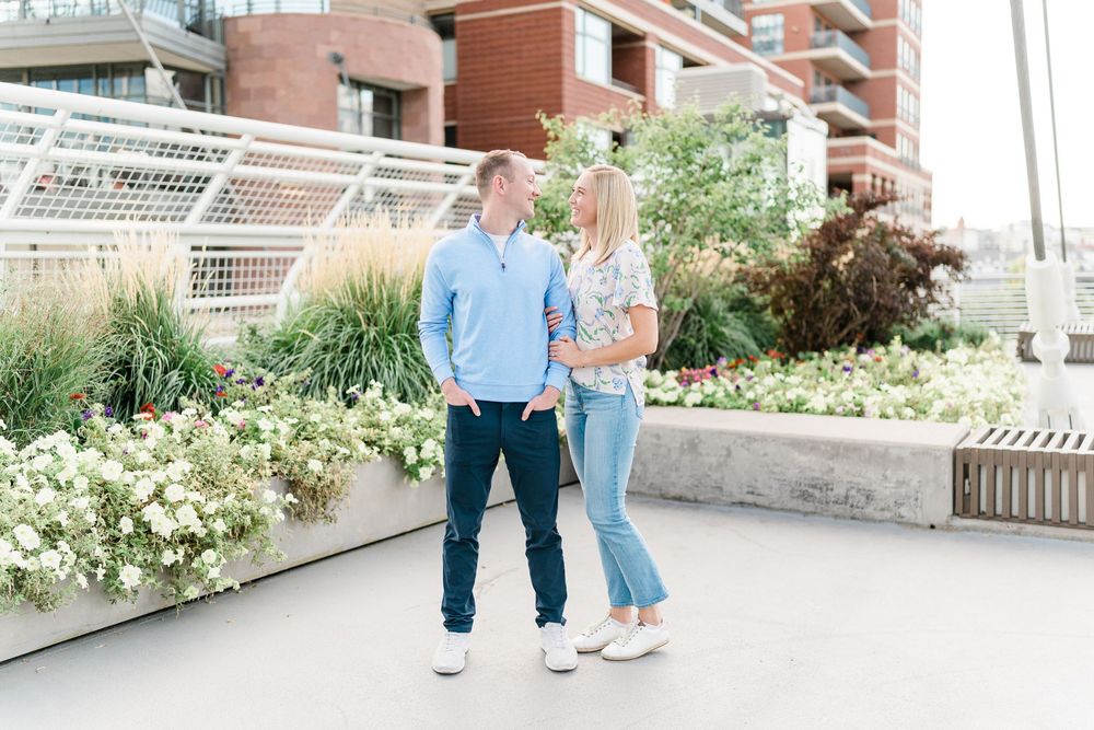 A couple stands together in casual attire on a walkway with flowering plants and modern architecture in the background.