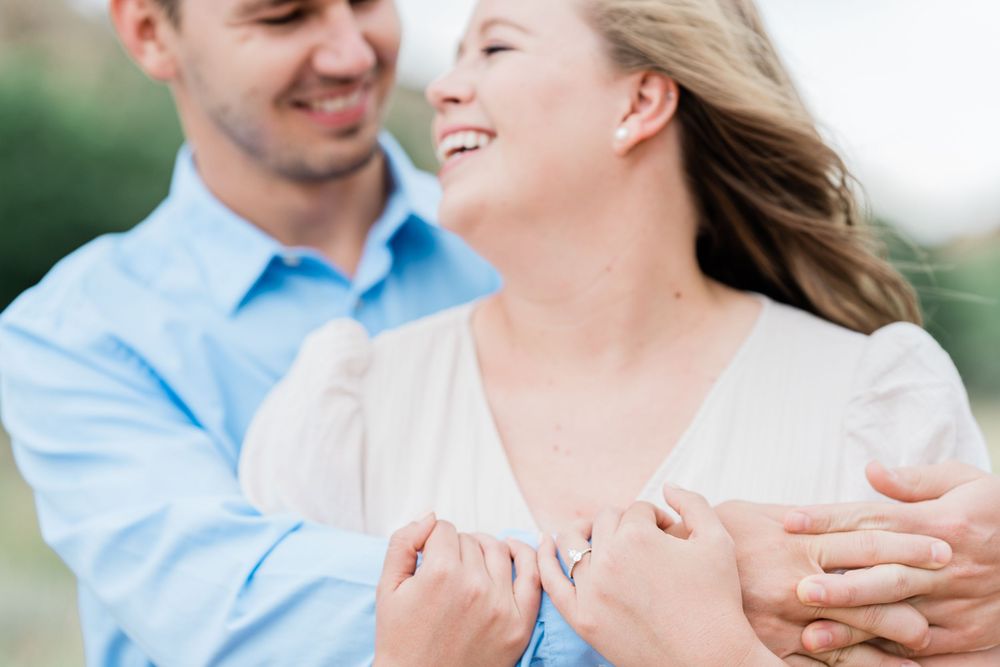 A loving couple embraces outdoors while laughing together, with her white sweater and his blue shirt against a soft background.