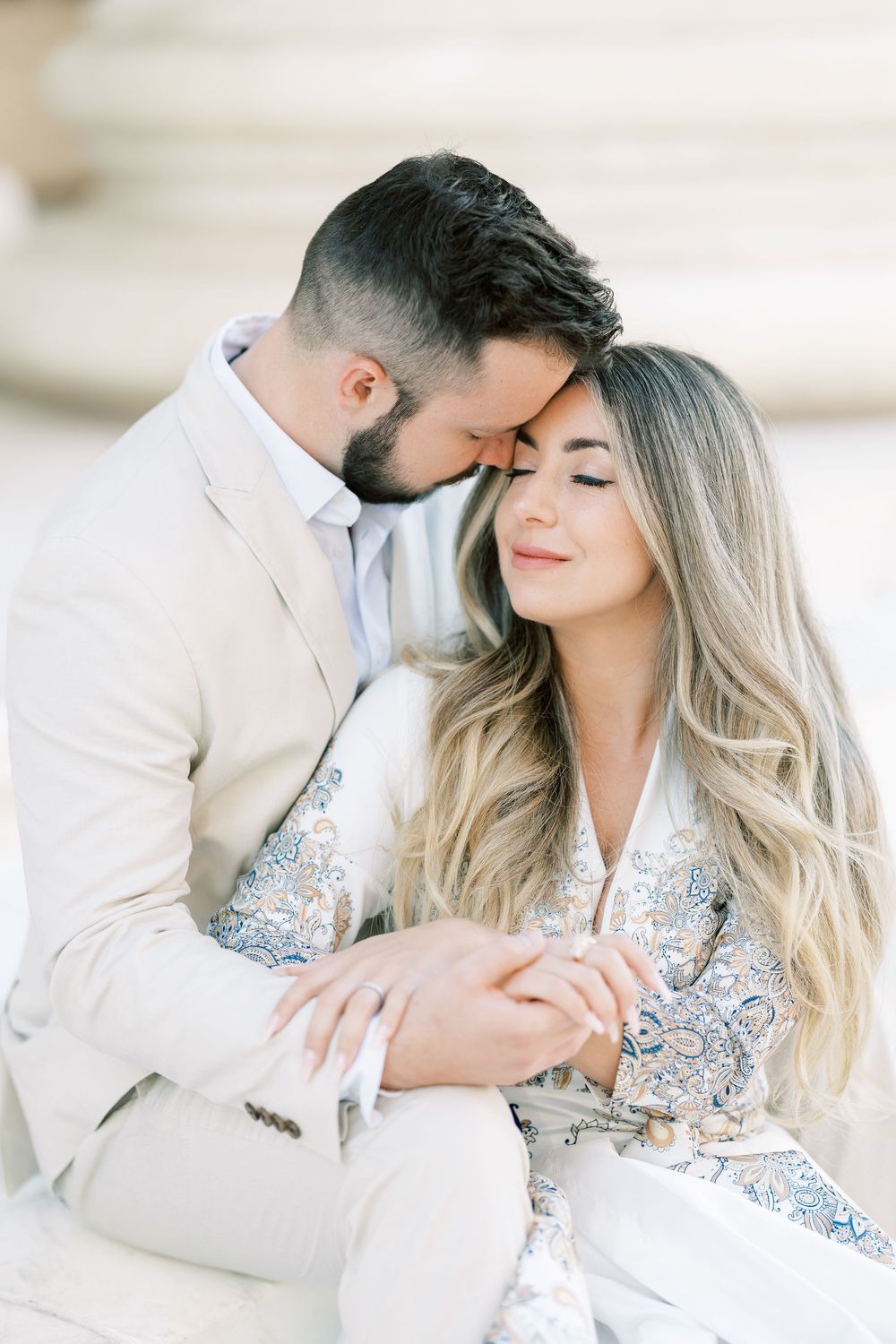 A couple shares an intimate moment while wearing elegant cream and floral attire against a bright neutral background.