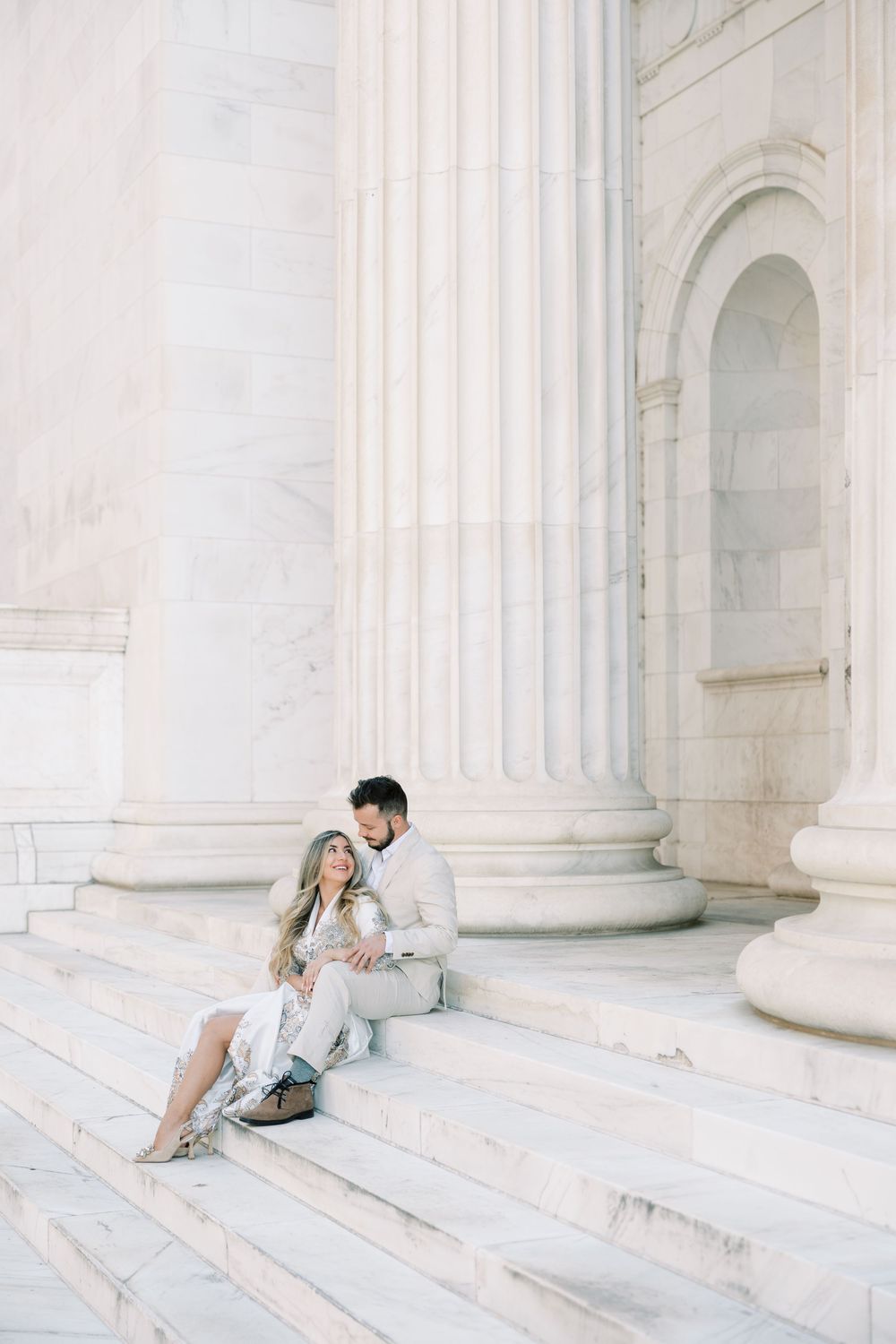 Couple in light colored clothing sits together on white marble steps against classic architectural columns.