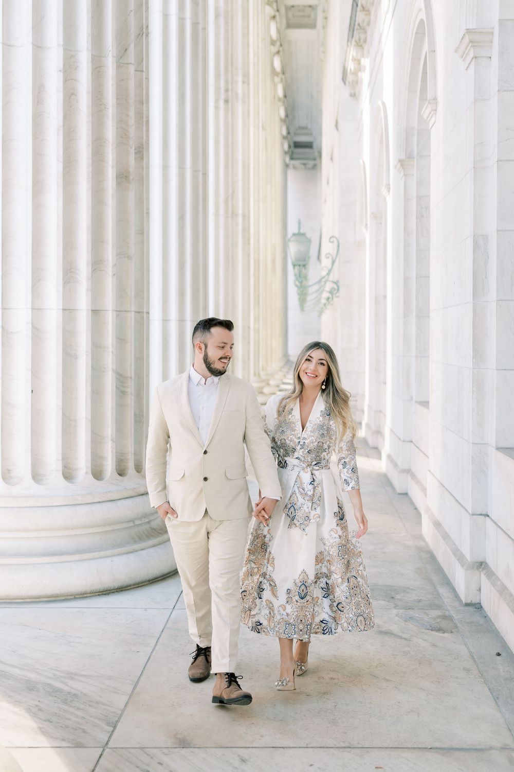 A couple in coordinated beige and white outfits poses along an elegant colonnade with white pillars and soft natural lighting.