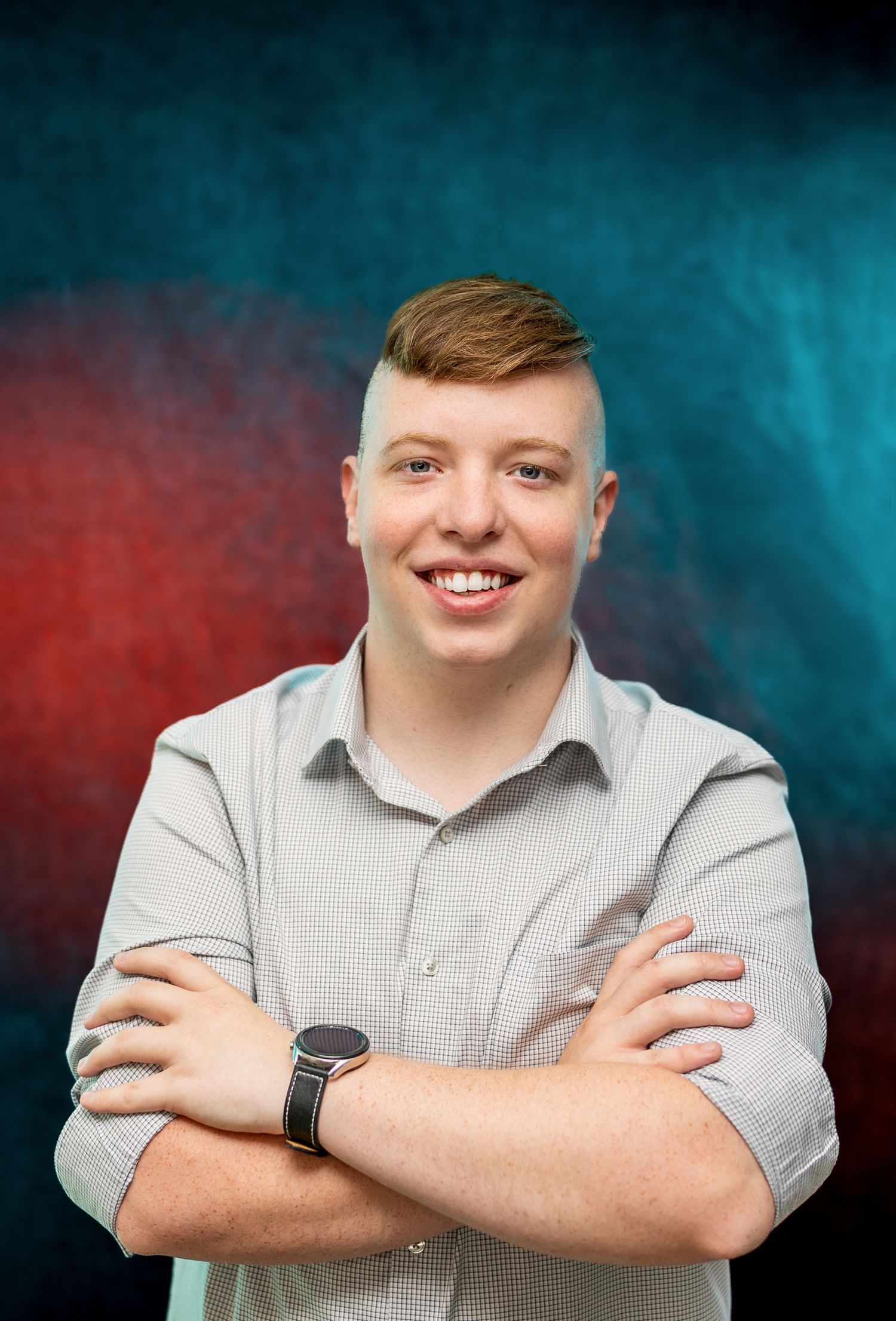 Smiling young man with short, styled hair and a light gray button-up shirt, standing with arms crossed in front of a red and teal gradient