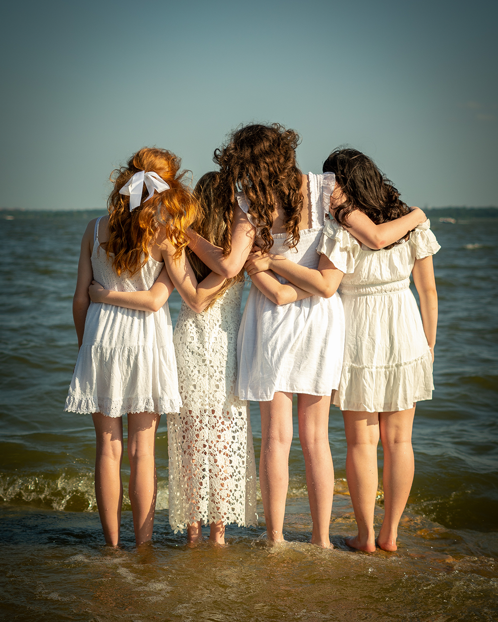Group of teenage females in a horizontal row on a beach, arms around each other, looking out toward the water.  Photo by Brian Balloon