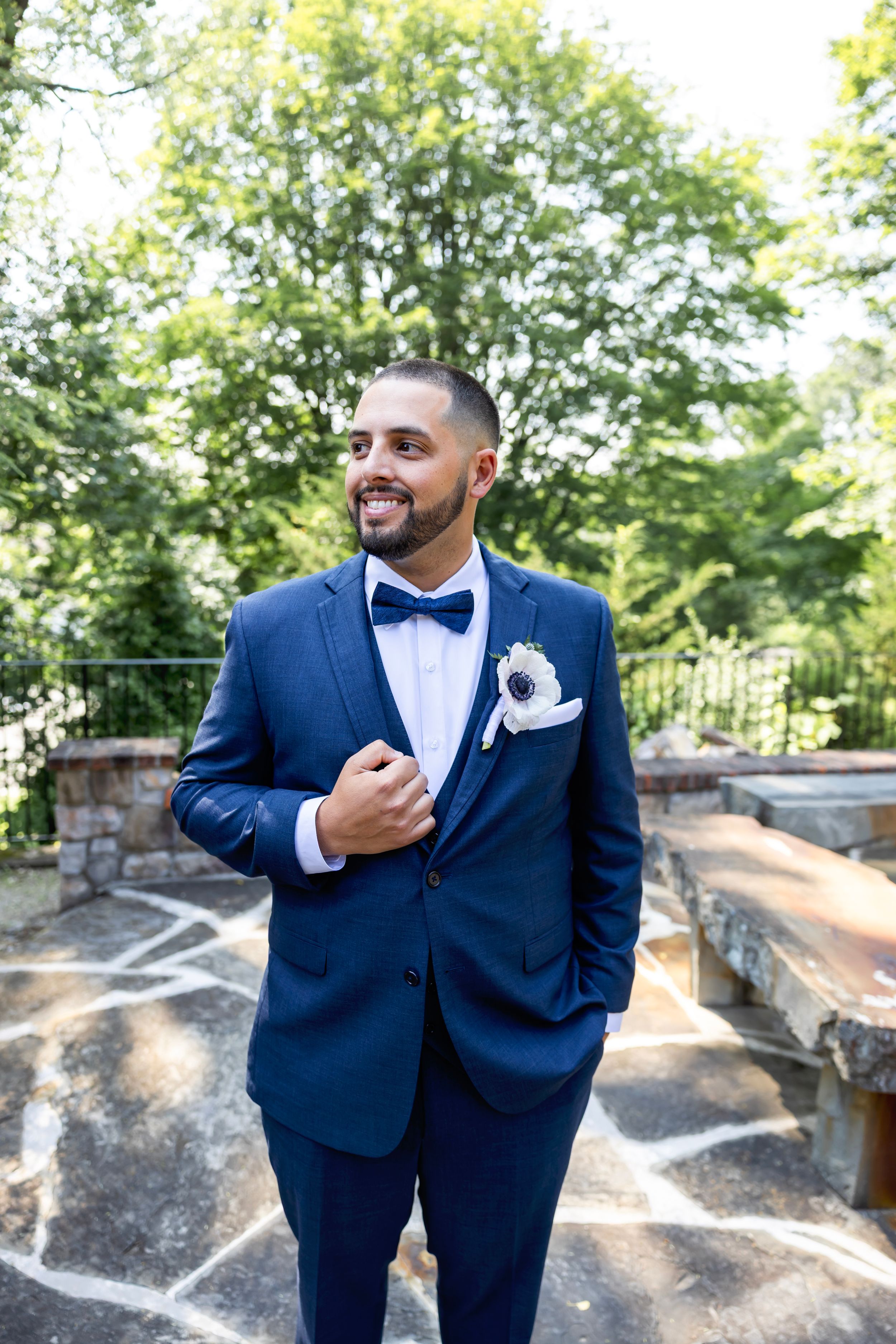 Groom in navy blue suit and bow tie poses outdoors near stone wall during wedding photoshoot.