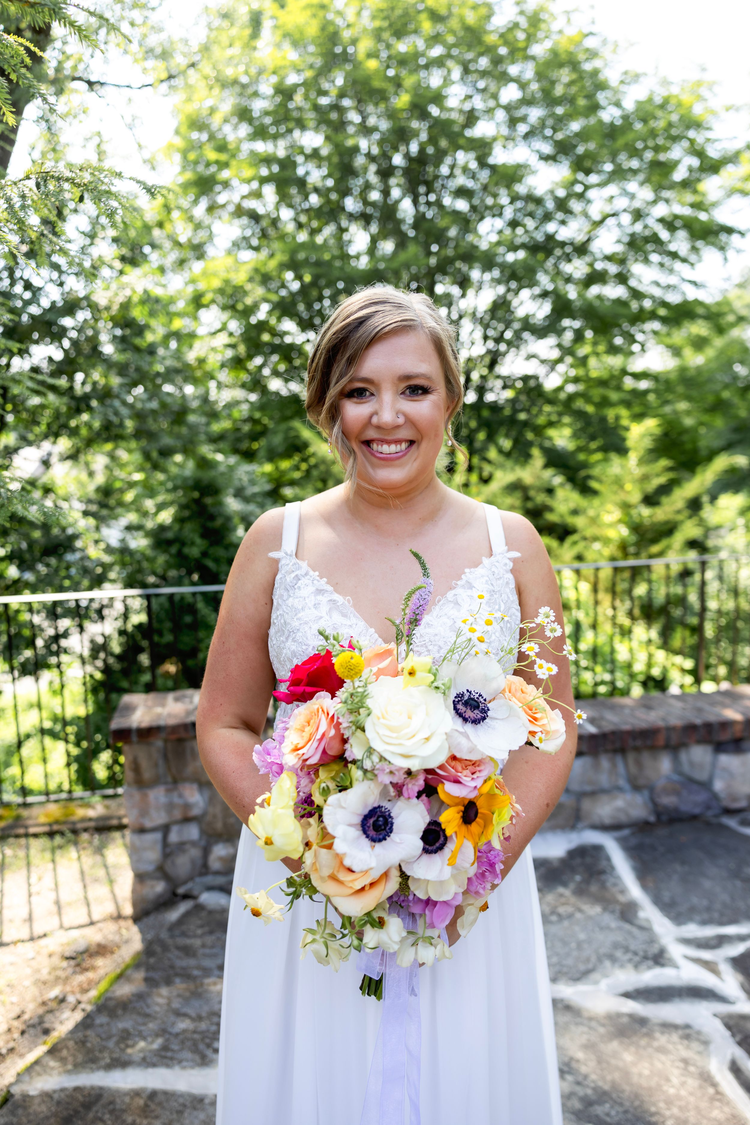 Bride in white wedding dress holds colorful bouquet with anemones, roses and sunflowers on stone patio with greenery backdrop.