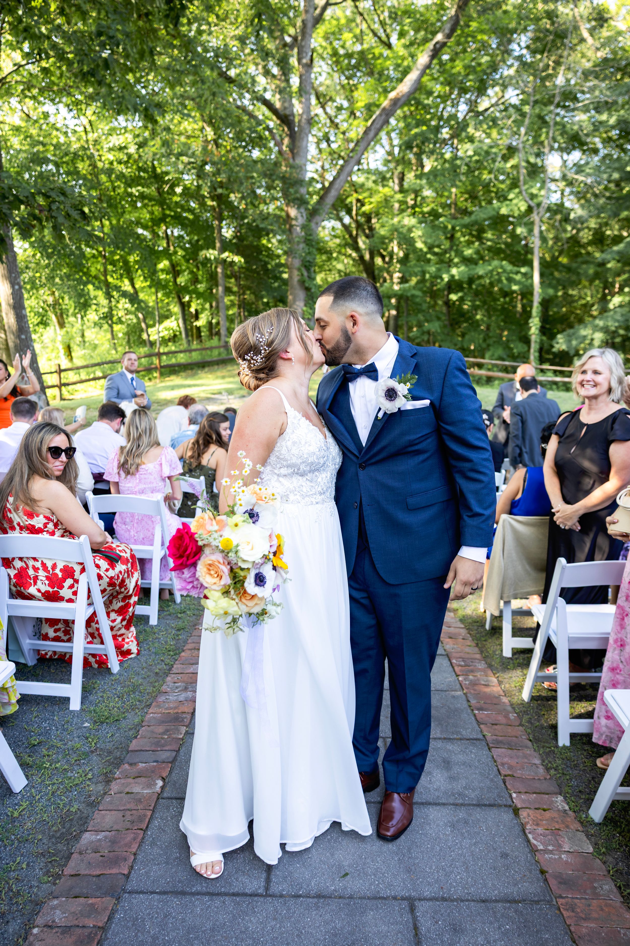 Wedding guests gather along a brick pathway surrounded by lush greenery during an outdoor celebration.