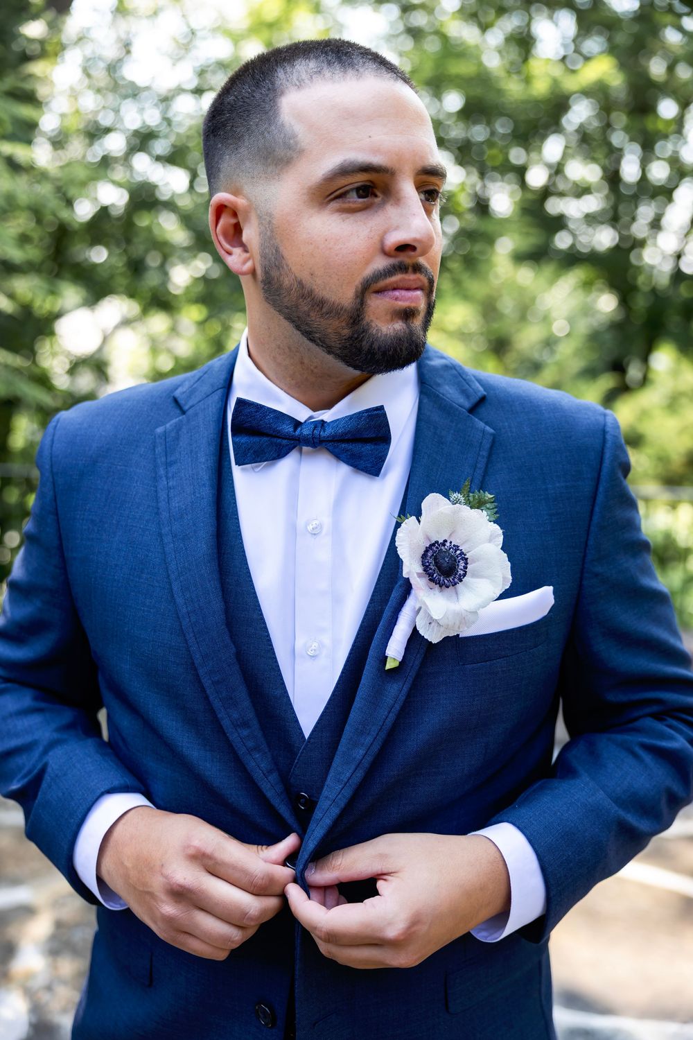 Close-up shots of groom in navy blue suit adjusting boutonniere and bow tie outdoors at wedding ceremony.