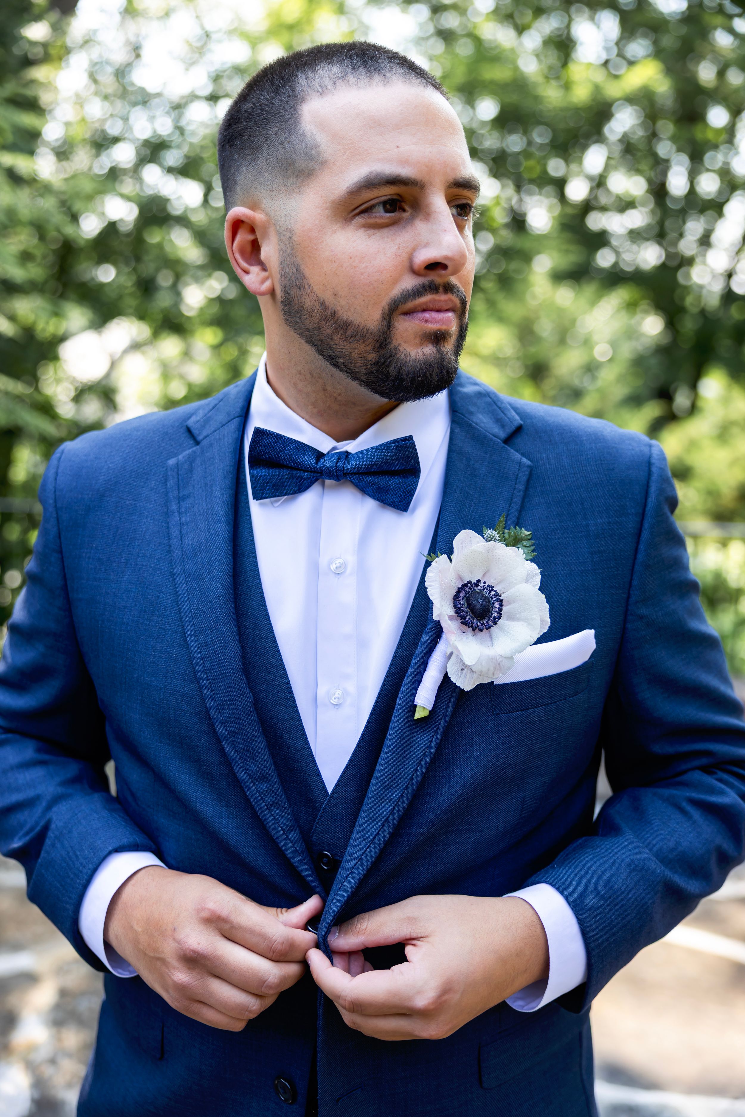 Close-up shots of groom in navy blue suit adjusting boutonniere and bow tie outdoors at wedding ceremony.
