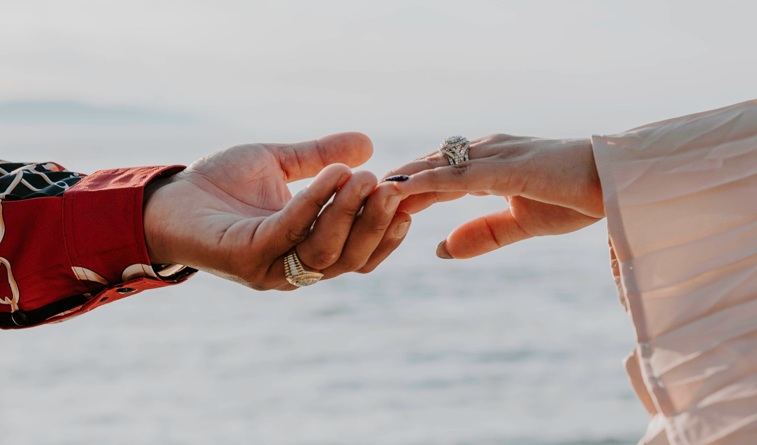 Two hands touching fingertips near the ocean, one wearing a diamond ring, symbolizing a romantic proposal.