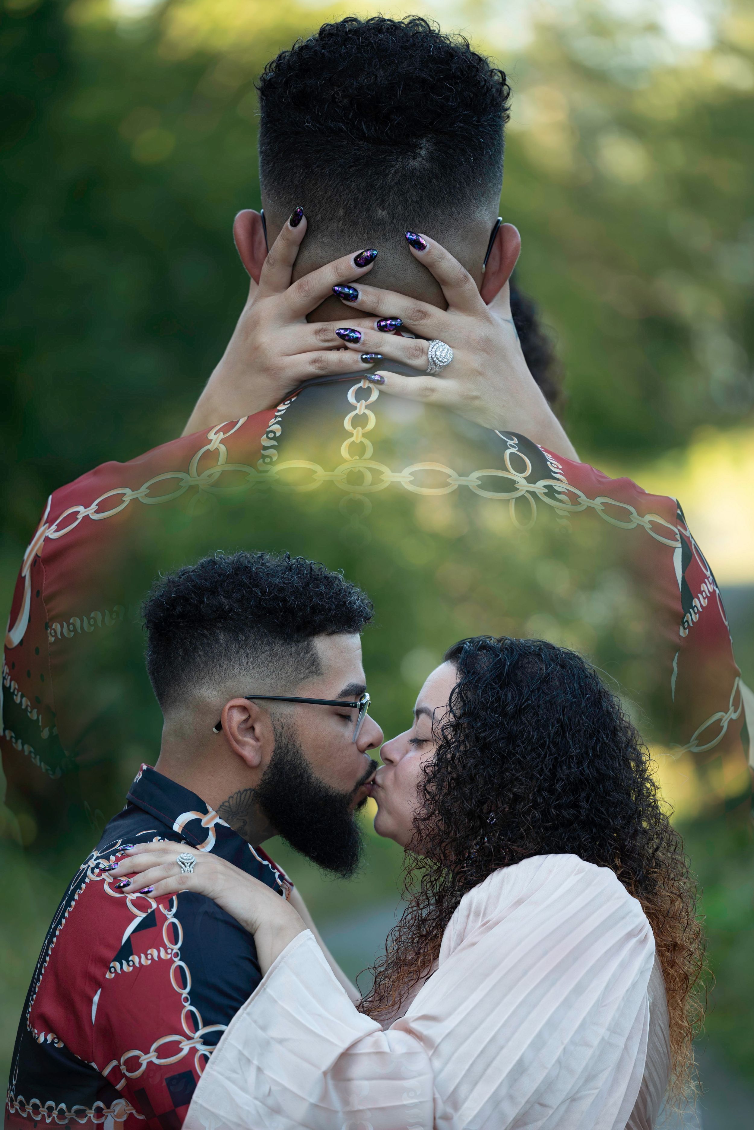 Couple sharing a romantic kiss outdoors, woman with curly hair and dark nails embracing bearded man in red chain-print shirt.