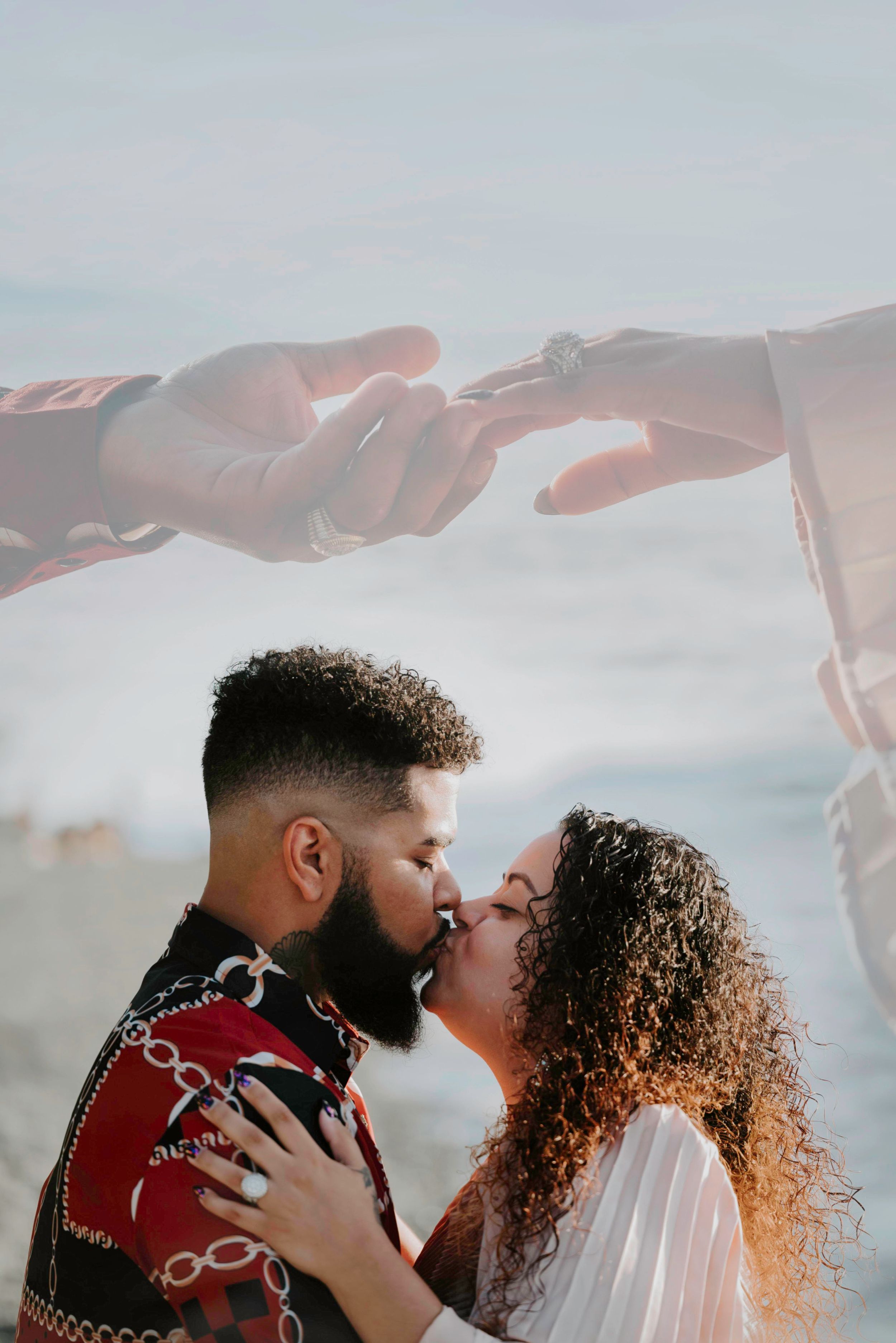 Couple sharing a romantic kiss on the beach with a double exposure of hands exchanging a ring overhead.