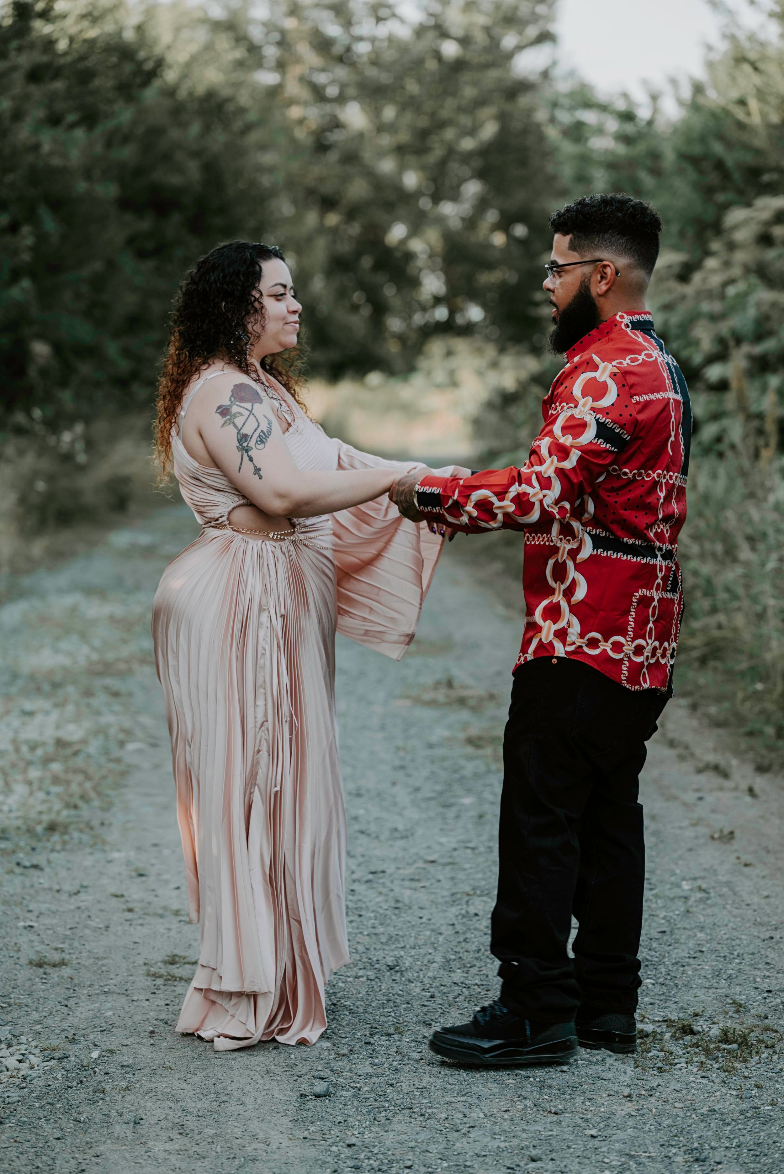 Couple holding hands on a dirt path, woman in pink pleated dress with tattoos and man in red chain-print shirt.