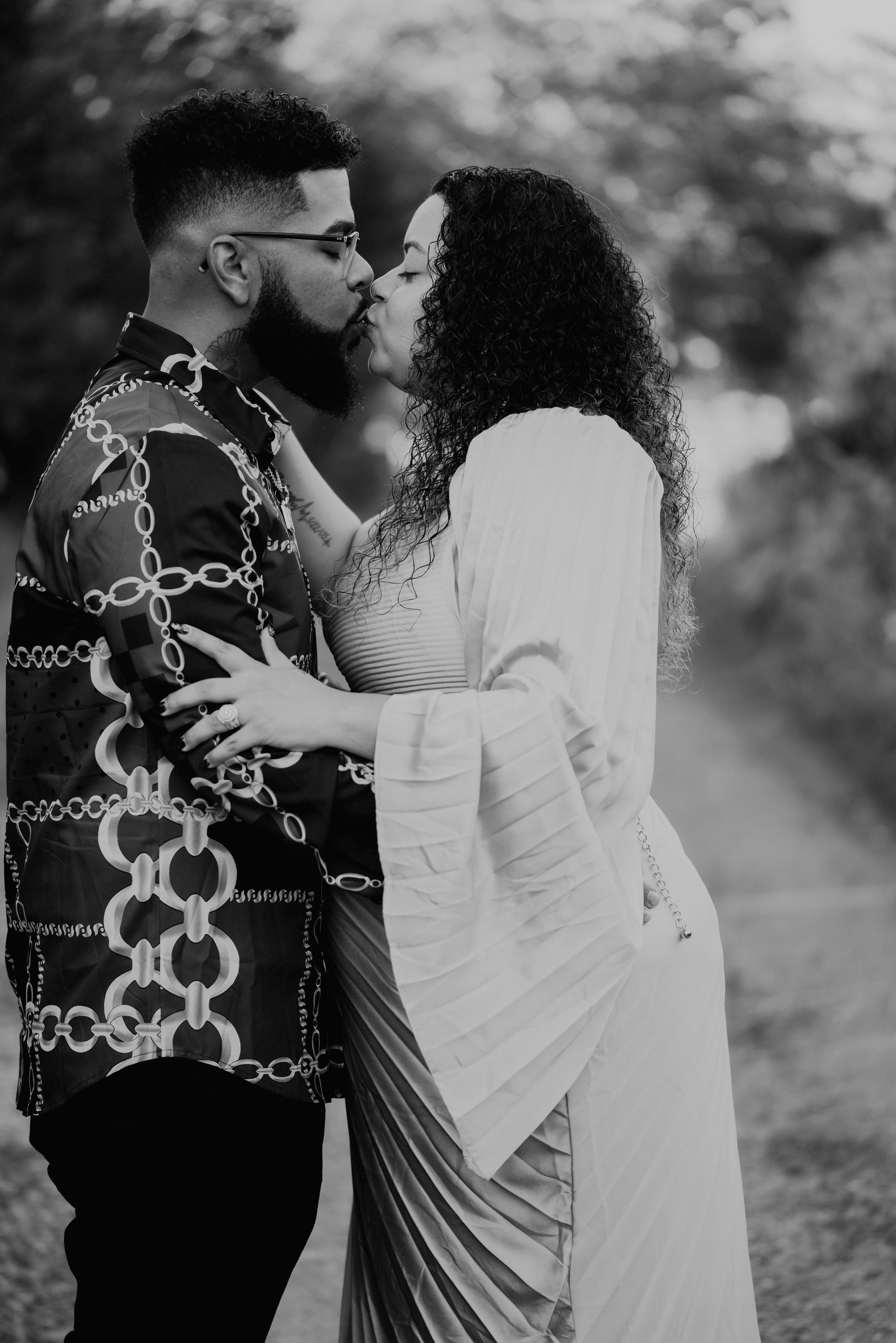 Black and white photo of a couple sharing a romantic kiss outdoors, woman in white dress, man in patterned jacket.