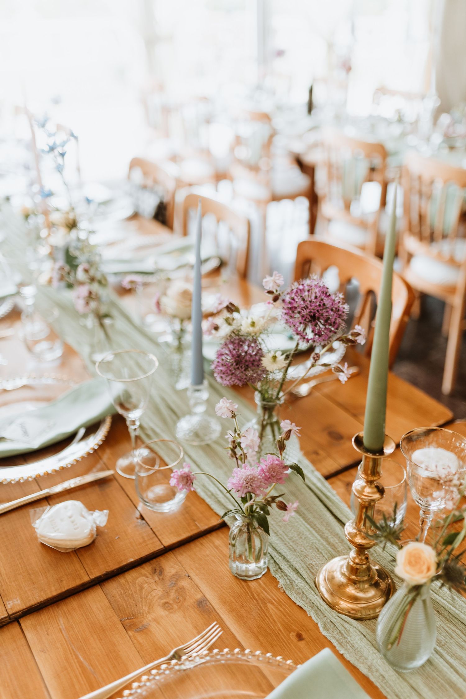 Rustic wedding table setting with pink wildflowers, sage green runner, and gold taper candles on wooden farmhouse table.