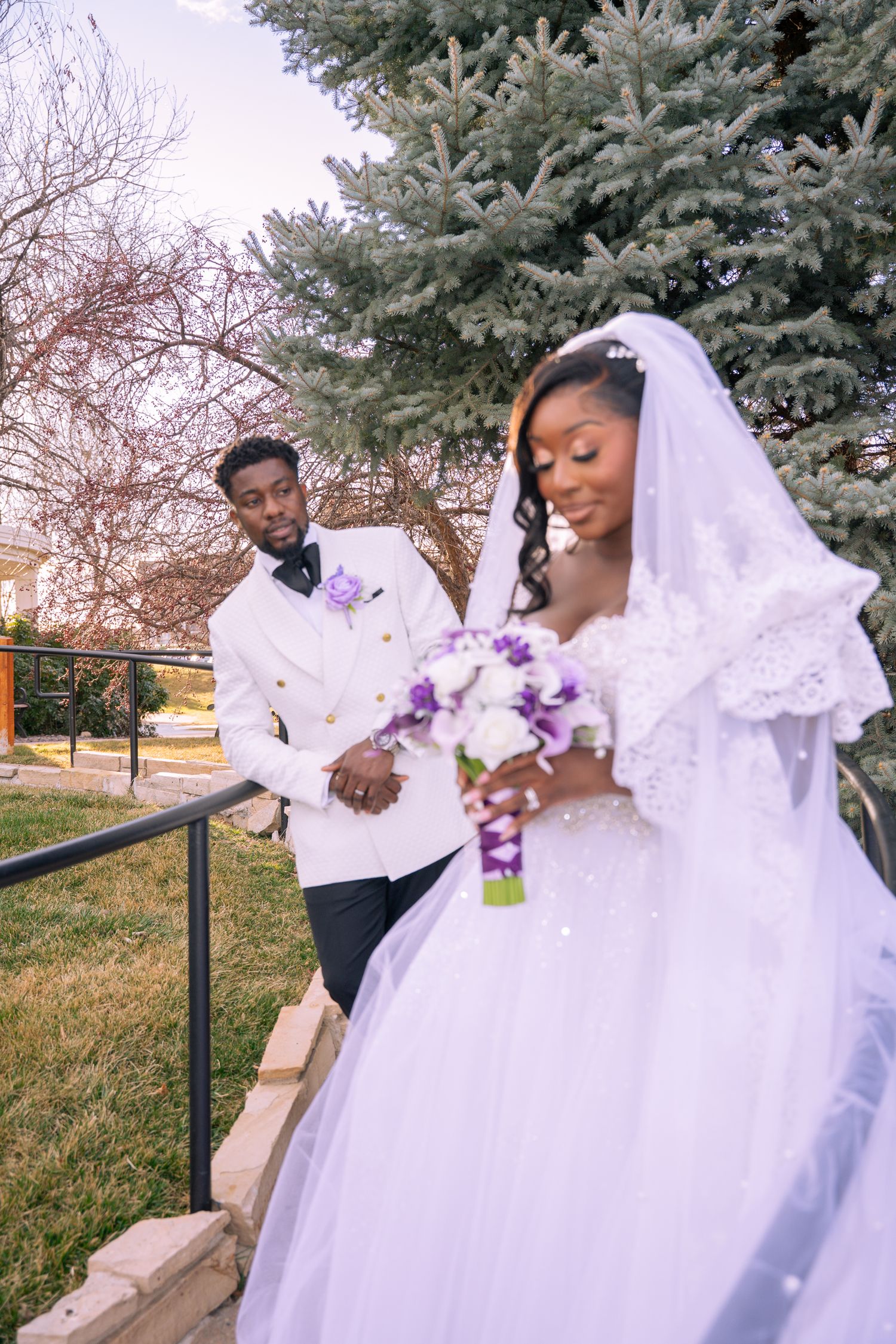 Newlyweds in white wedding attire sit together outdoors with a purple bouquet on a sunny day.