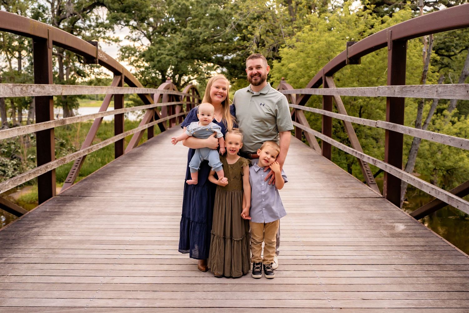 Family of four standing together on a wooden bridge in a park setting with green trees in the background.