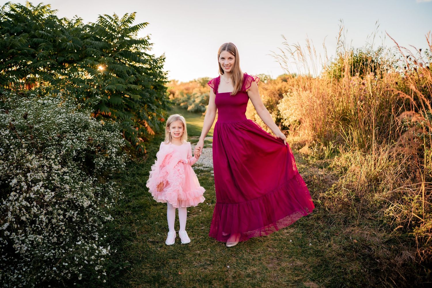 A burgundy gown and pink tulle dress twirl in golden sunlight among tall grasses and greenery at sunset.