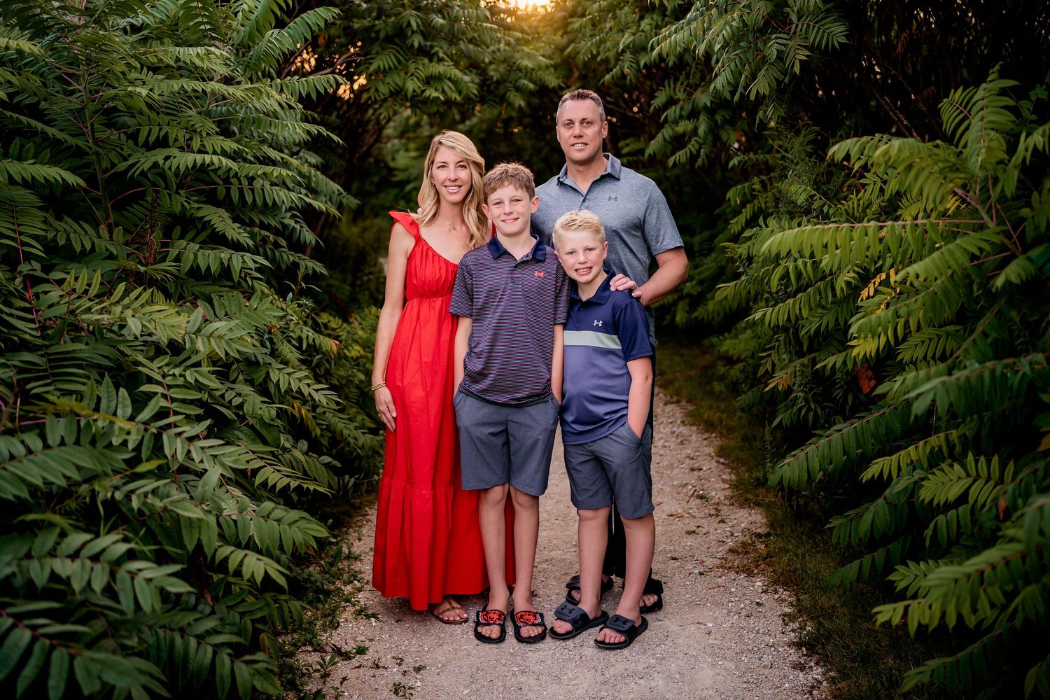 A family of four stands together on a garden path surrounded by lush ferns at sunset.
