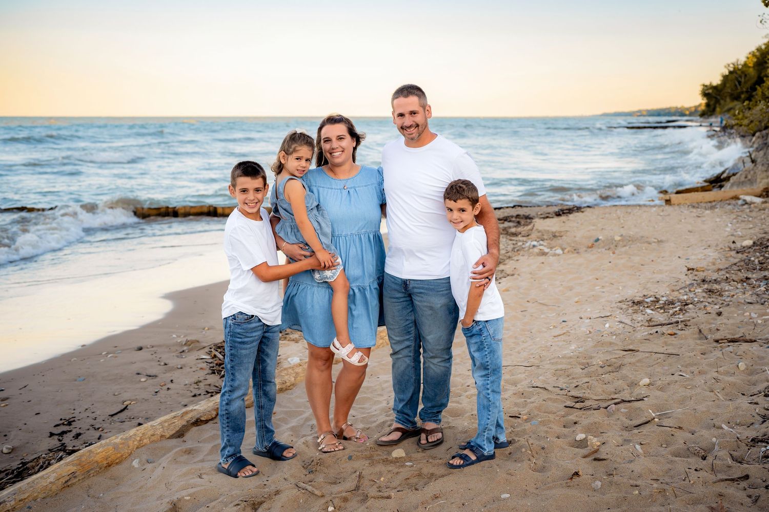 A group of five people stand together smiling on a beach at sunset with waves and coastline in the background.