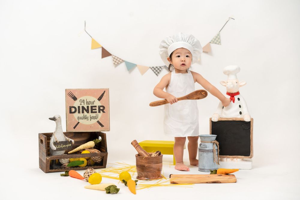 Child in chef outfit plays with toy kitchen set and cooking accessories against white background.