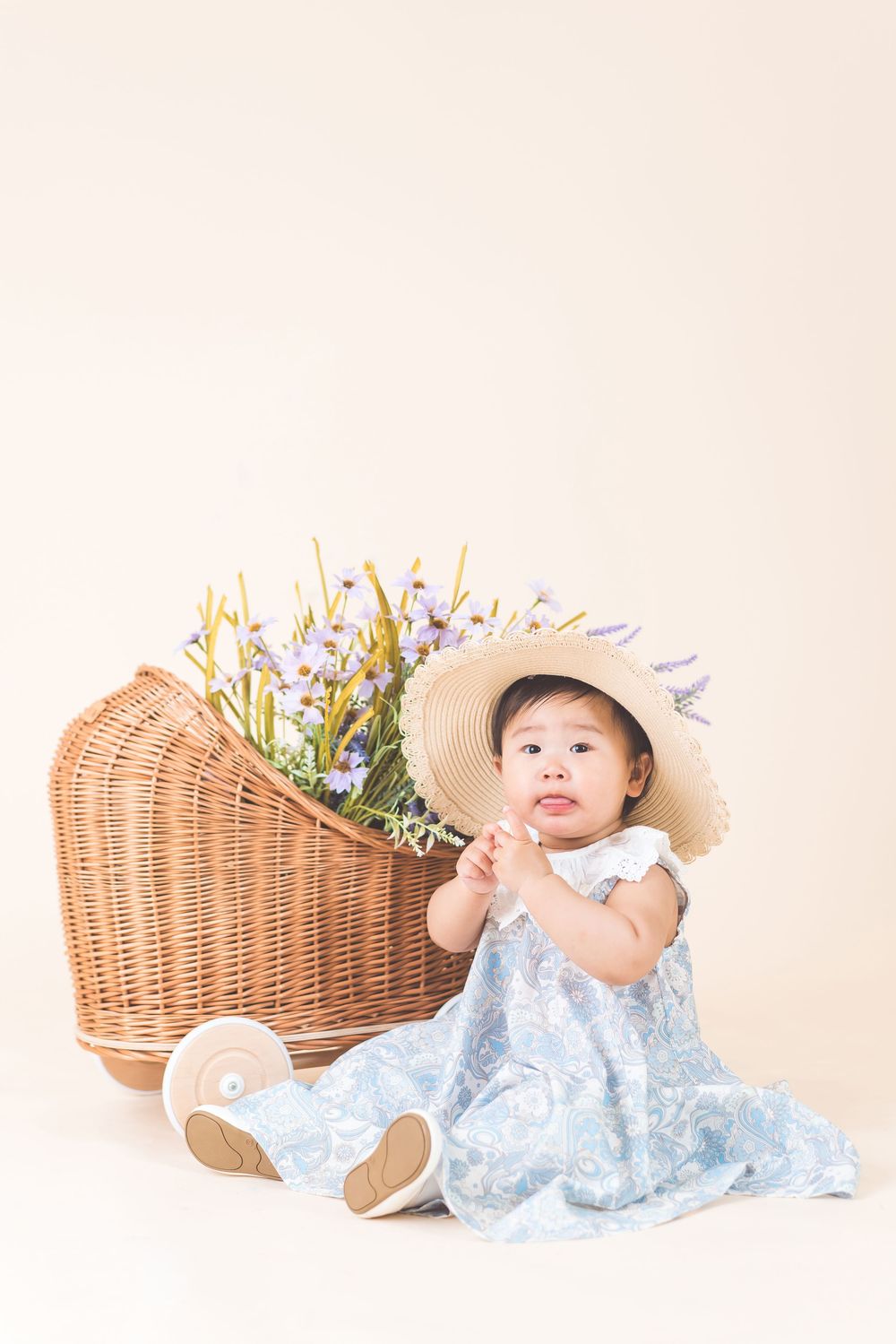 Baby in blue dress and sun hat sits next to wicker basket filled with spring flowers.
