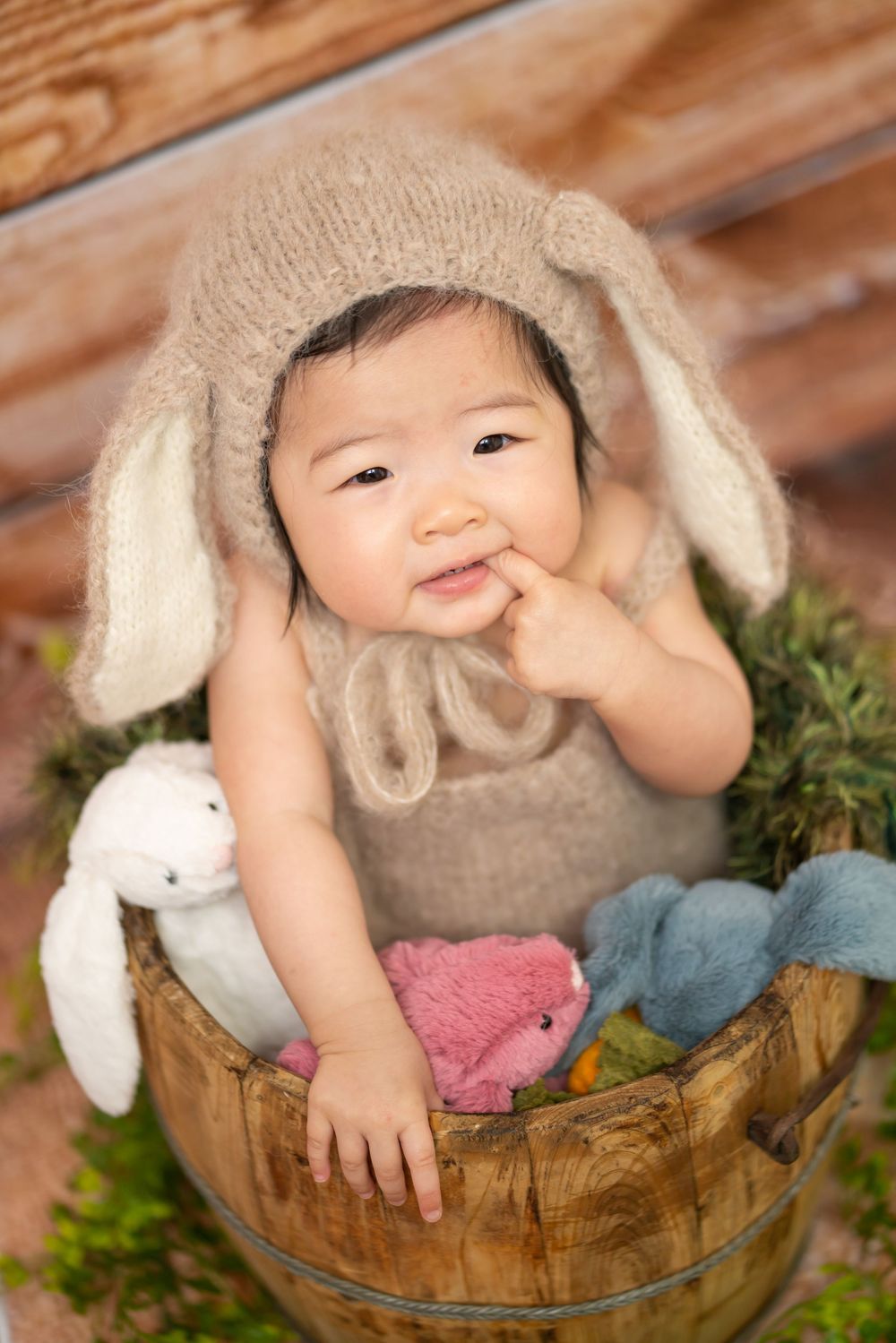 Baby in knit bunny hat sitting in rustic wooden bucket with stuffed animals.