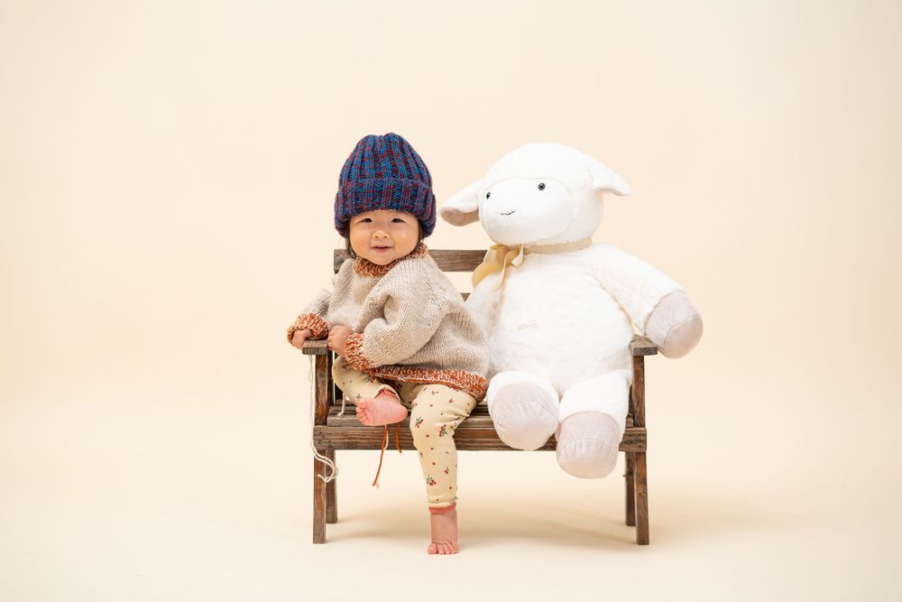 Child in knit hat sitting with plush lamb toy on wooden bench.