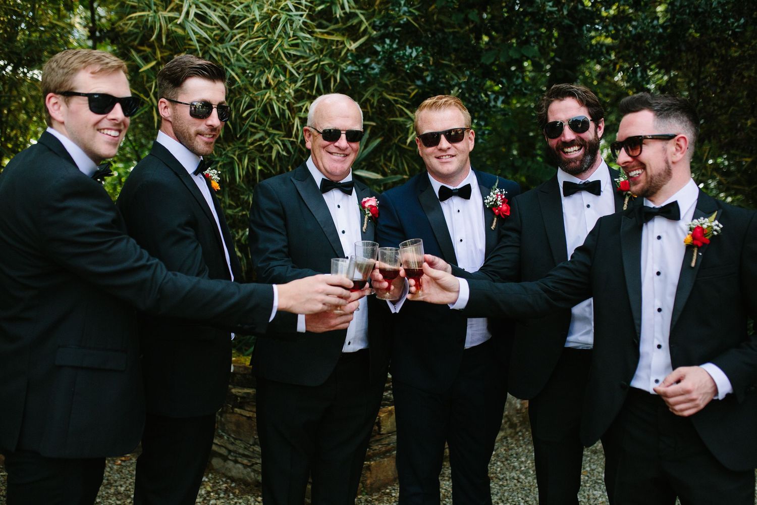 Groomsmen in black tuxedos and sunglasses toasting drinks together at outdoor wedding celebration.