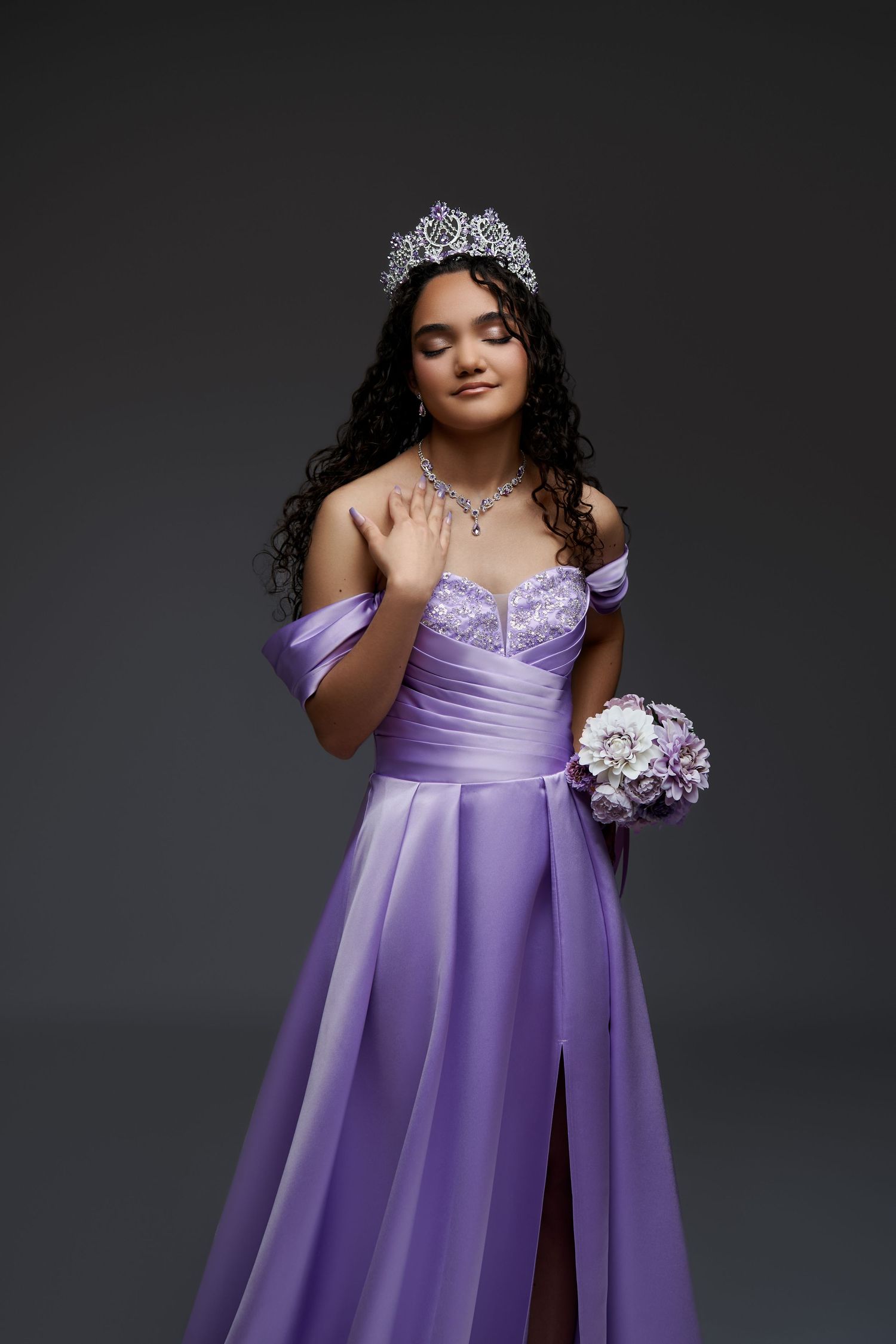 Beautiful quinceañera gown in lavender satin features off-shoulder design, crystal tiara and bouquet against a backdrop. gricel photography