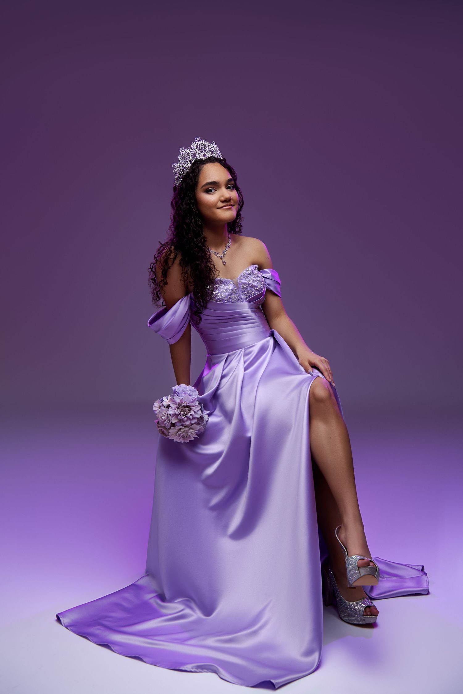 A stunning lilac off-shoulder formal gown with a leg slit, styled with a tiara and matching bouquet against a purple backdrop. sweet sixteen