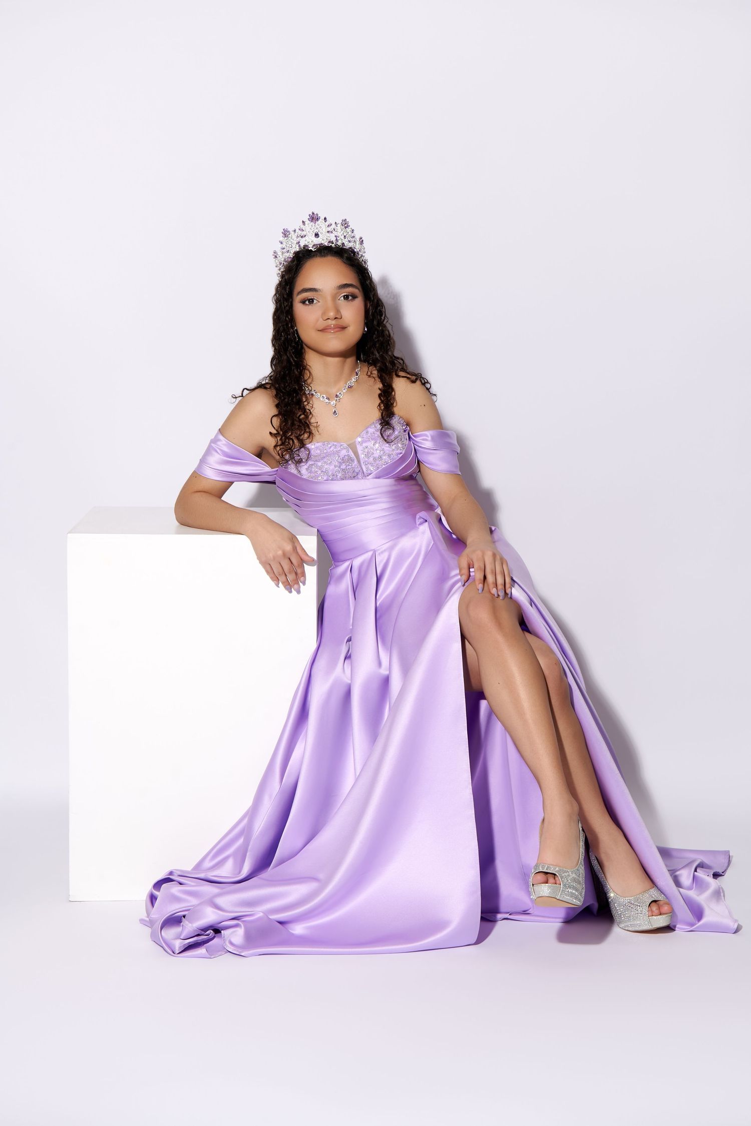A person in a lavender off-shoulder formal gown with a tiara sits elegantly against a white background with legs crossed. NJ photographer