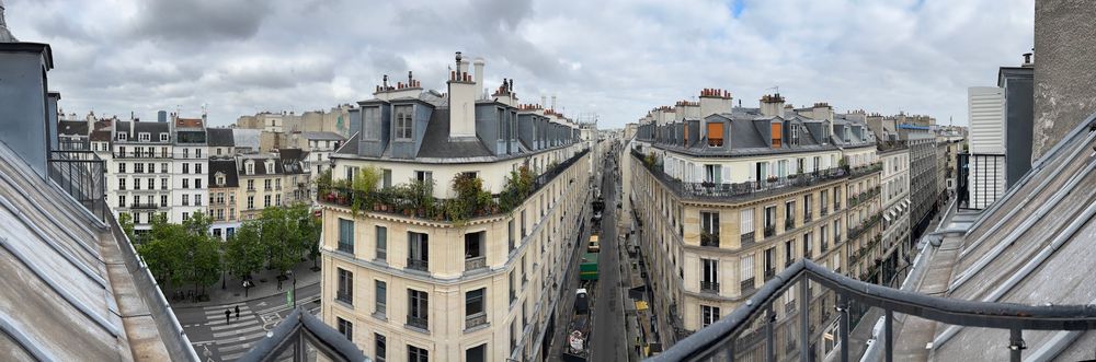 A scenic aerial panorama of Paris rooftops with traditional Haussmann architecture and chimneys under cloudy skies.