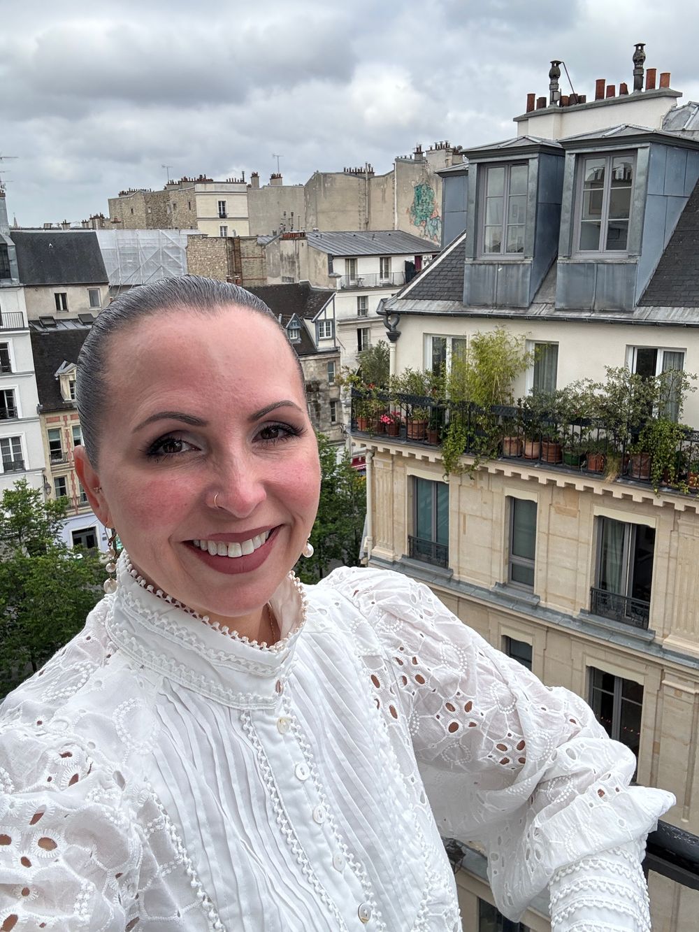 Woman in white lace dress stands on balcony overlooking Parisian rooftops against cloudy sky.