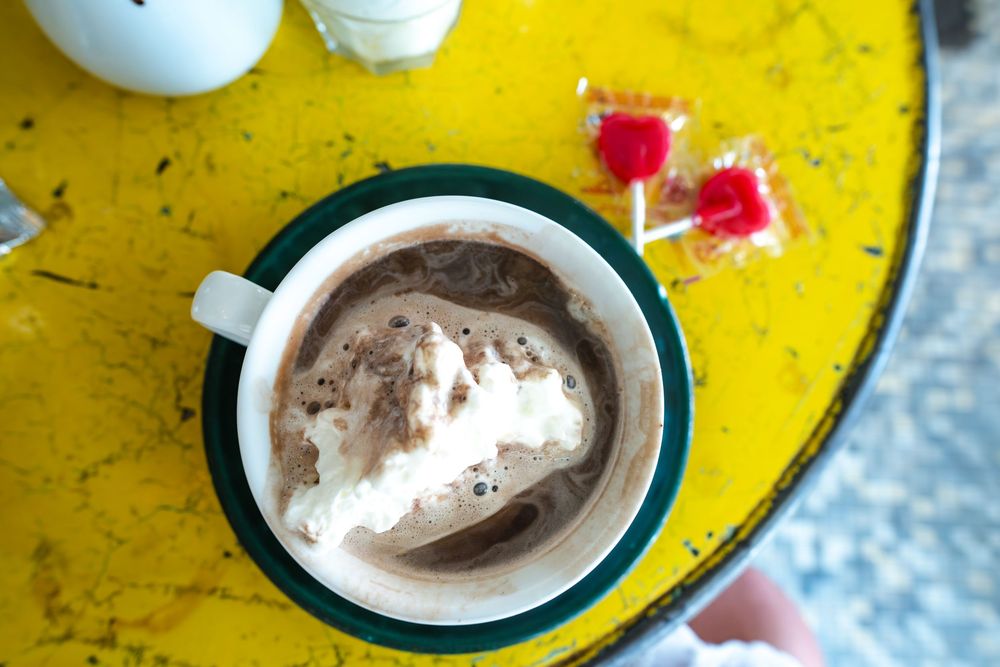 A cup of hot chocolate with whipped cream and cocoa powder on a bright yellow surface with pink candy wrappers nearby.