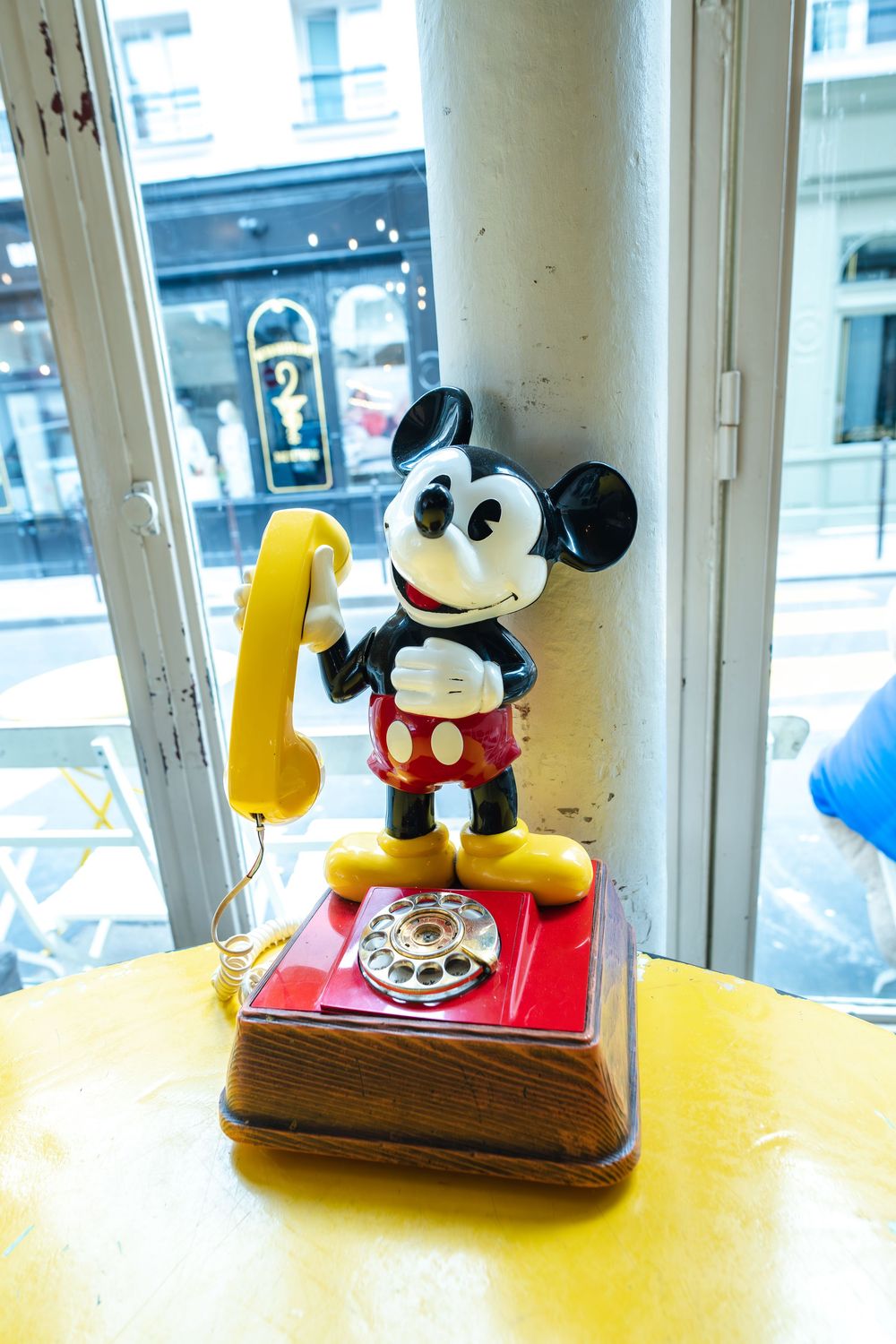 Vintage Mickey Mouse rotary telephone with yellow handset sits on wooden base near window in quaint setting.