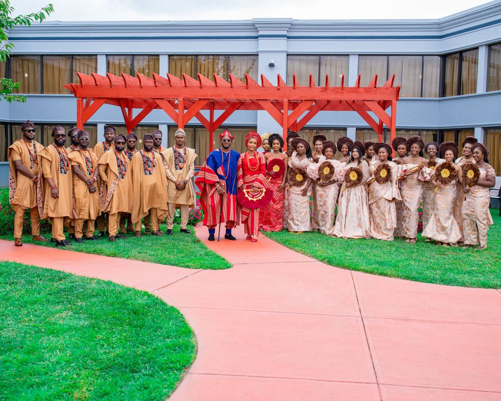 Wedding party members in vibrant red and beige attire pose together under a red pergola on a manicured lawn.