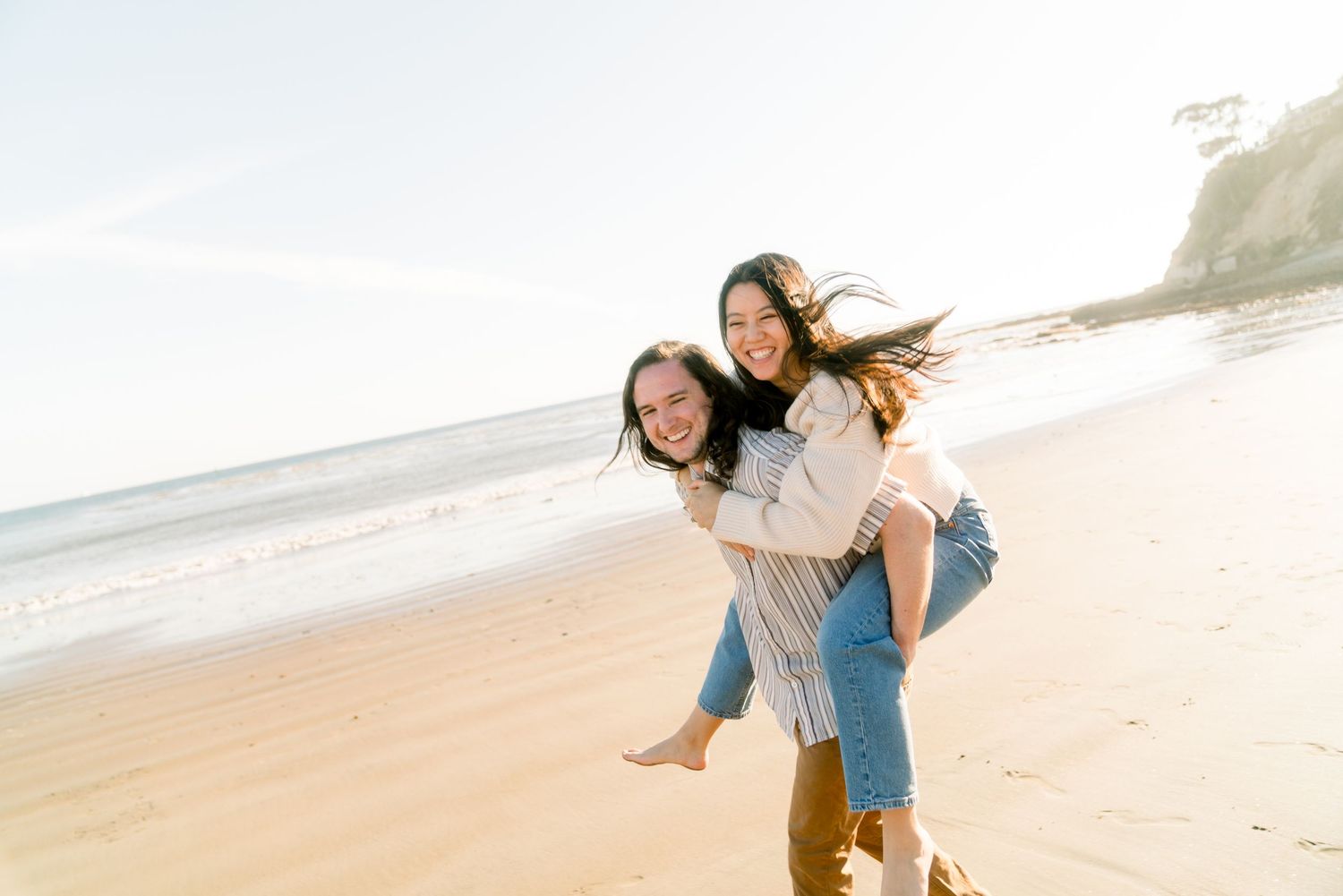 Two friends sharing a fun piggyback ride while enjoying a sunny day at the beach with cliffs in the background.