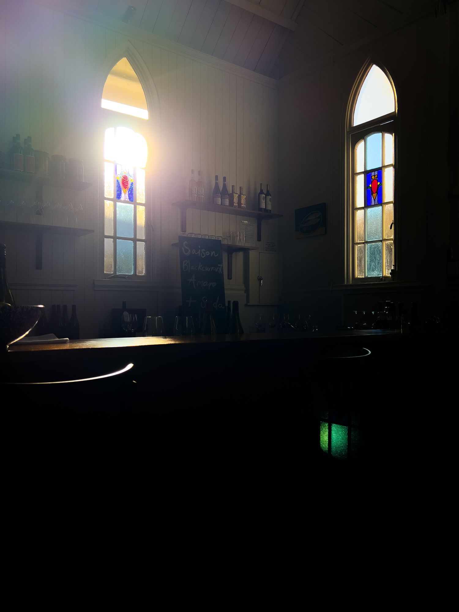 Sunlight streams through two arched stained glass windows in a dimly lit church sanctuary with bottles in the background.