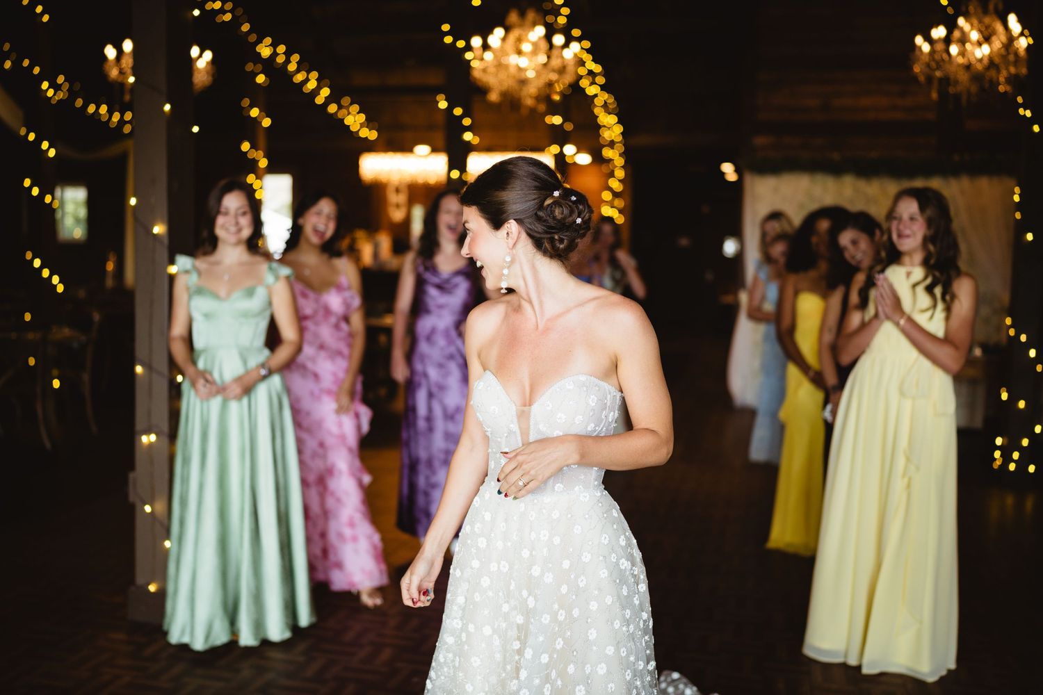 A group of wedding guests dance and celebrate on a dimly lit dance floor with twinkling lights in the background.