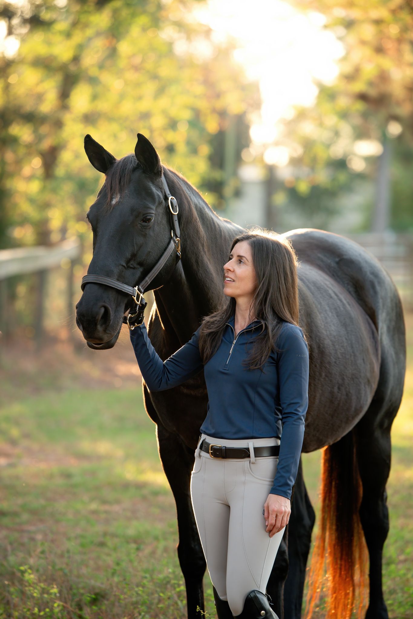 Equestrian in blue sweater and riding pants stands beside elegant black horse in autumn setting at sunset.