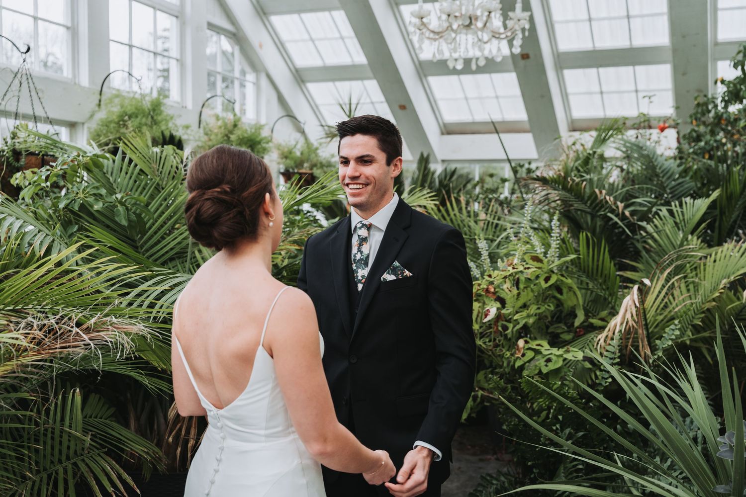 Two people in wedding attire share a moment together inside a greenhouse filled with lush tropical plants and palms.