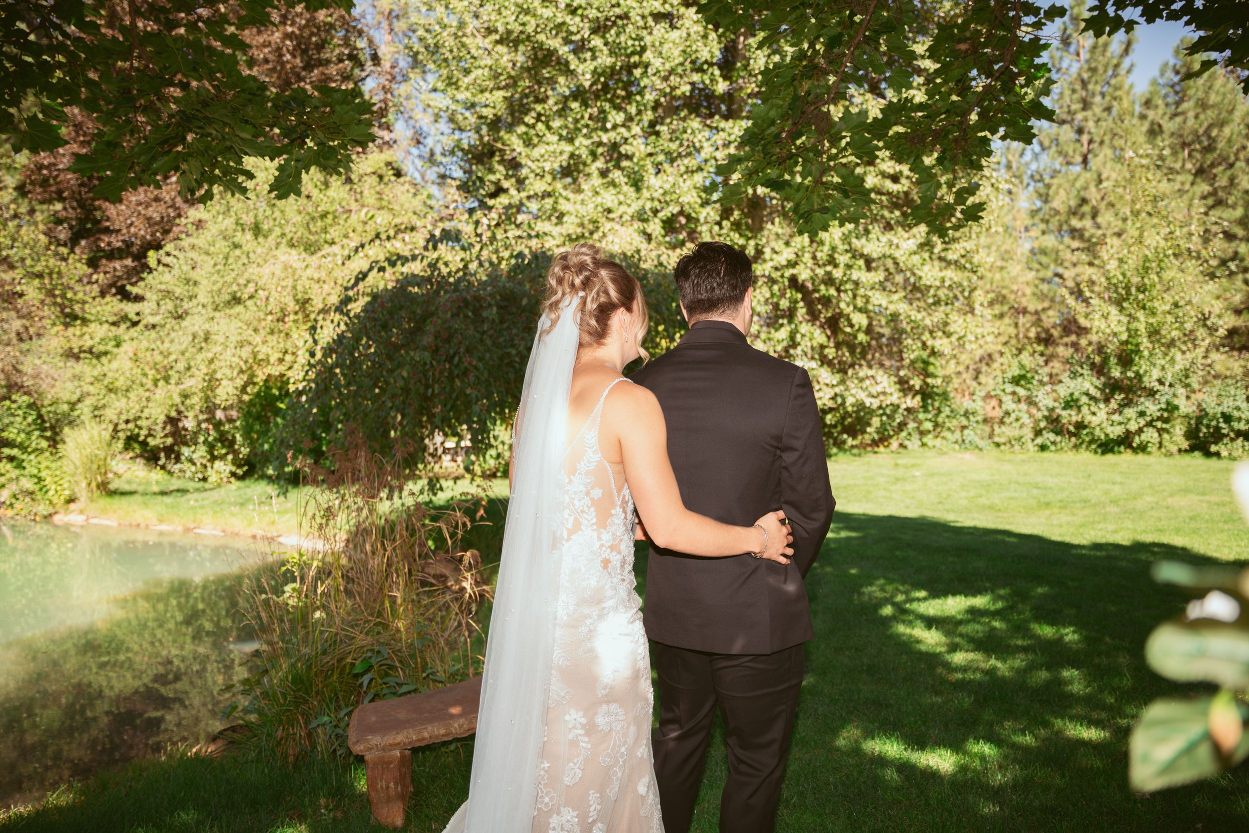 A couple in formal wedding attire stands together in a sunlit garden surrounded by lush green foliage.